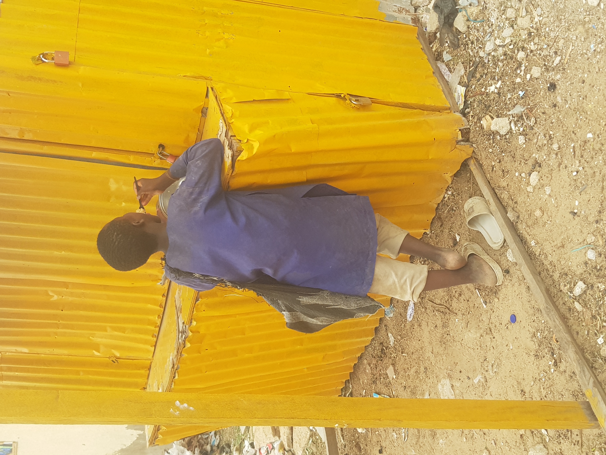A young person paints a yellow metal shack in Dabawa, Nigeria. The person is wearing a blue shirt and beige pants. One sandal is next to the person's feet on the dirt ground. Debris is visible.