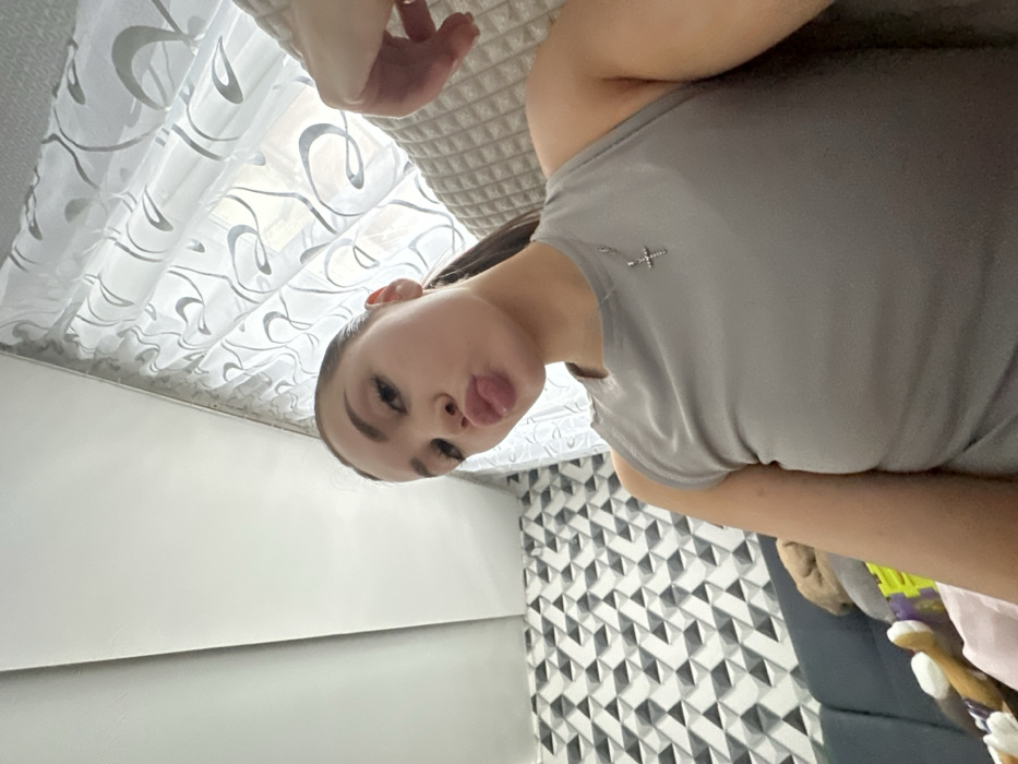 A young adult female is depicted in a close-up, upward-angled view, situated indoors within what appears to be a residential setting in the city of Chaikovsky, Russia. The subject, with dark hair pulled back, is making a "duck face" expression, puckering her lips forward with the tip of her tongue slightly visible. She is wearing a light gray, sleeveless top, and a silver-colored necklace with a small, jeweled cross pendant rests on her chest. Her right forearm and hand are partially visible on the right side of the frame, resting on a beige, textured surface.
The background includes a light-colored, possibly white, ceiling that slopes slightly upward on the left. To the subject's left, a wall is covered with a repeating geometric pattern of three-dimensional triangular or pyramidal shapes in shades of gray and white. Below this wall section, a dark-colored sofa or couch is partially visible, with indistinct objects, including what appears to be a light brown plush item, resting on it. To the subject's right and behind her, a window is covered by a sheer white curtain featuring a repeating gray, swirling or floral design. The scene is illuminated by ambient light, possibly combining natural light from the window with indoor lighting. No other individuals are present or interacting with the subject.