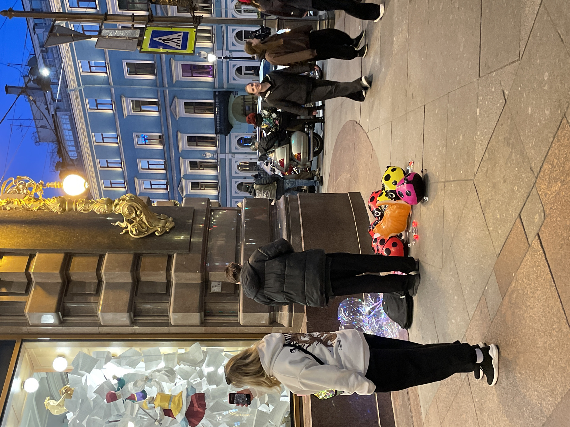 This evening street scene in Saint Petersburg, Russia, depicts a sidewalk teeming with activity under twilight skies. In the foreground, a blonde woman in a white hoodie and black pants is engrossed in her phone. Beside her, a vibrant display of several ladybug-themed balloons in red, yellow, and pink, along with a clear, sparkling balloon, rests on the paved ground.
Further back, a group of individuals gathers near a light-colored scooter or moped parked on the sidewalk. One person in a dark jacket smiles towards the camera, while others stand nearby. The background is dominated by a majestic, multi-story building painted a striking blue, featuring numerous well-lit windows. Its architectural style, including decorative cornices and window frames, is characteristic of Saint Petersburg.
To the left, an ornate golden street lamp casts a warm glow, adding to the city's historical aesthetic. Above the street, a blue and yellow pedestrian crossing sign is visible. The sky is a deep, dark blue, indicating either dusk or early night, with various artificial lights illuminating the street, building interiors, and a shop window display in the lower left that features abstract white shapes and colorful figures. People are dressed in jackets, suggesting cooler weather. The pavement is a mix of speckled gray and brown tiles. No discernible text is clearly visible on signs or objects within the image.