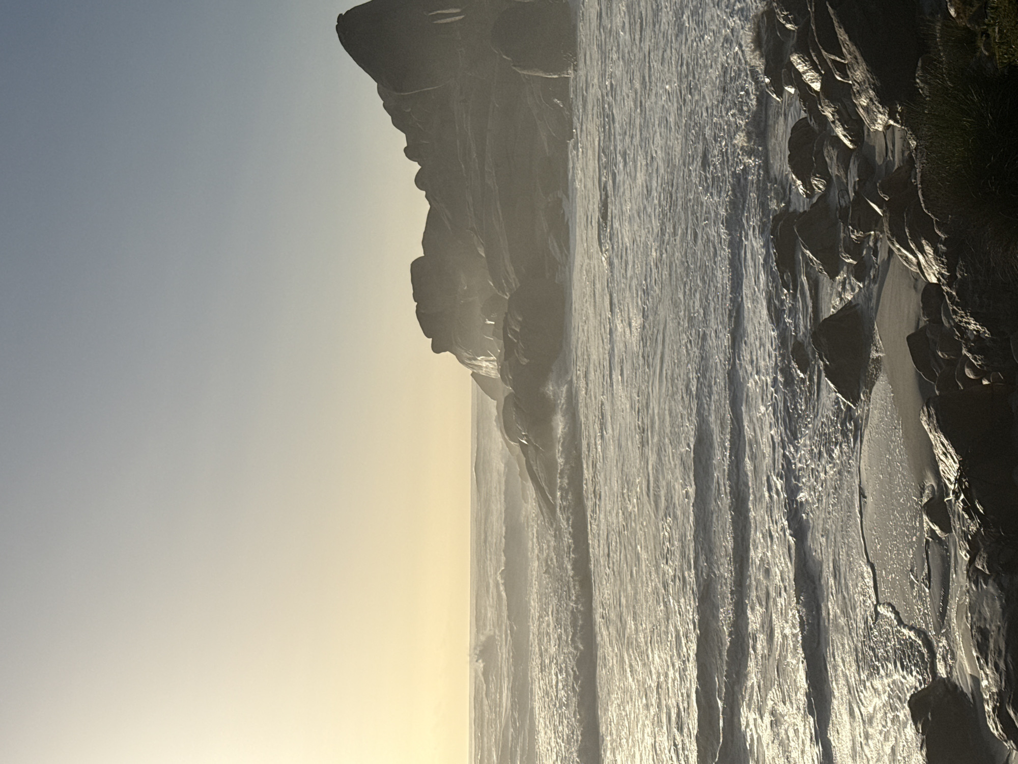 The image shows a rocky coastline at sunset. The location appears to be in or near Cape Town, South Africa. The sky is pale with a yellow tint from the setting sun. Tall, dark cliffs line the left side of the frame. Waves crash against the rocks and sand. Water streaks the beach. There are no people visible in the image.