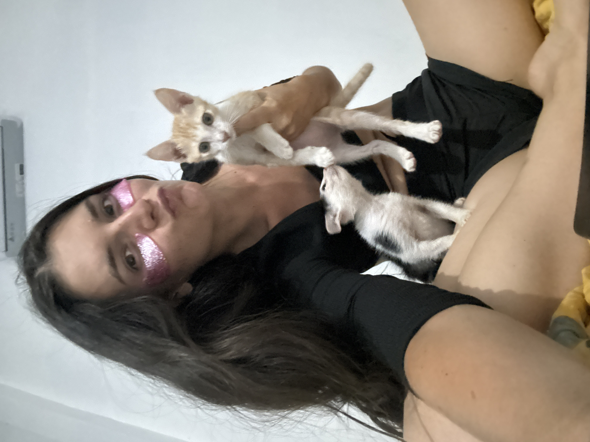 A woman with dark hair and glittery pink under-eye patches makes a pouty face while holding two kittens. One kitten, ginger and white, is held in her right arm, while a black and white kitten is positioned lower, near her lap. The woman is wearing a black, long-sleeved top and black shorts. The background is a plain, light-colored wall with an air conditioning unit mounted high up. The scene appears to be indoors, possibly a bedroom or living space. The overall lighting is soft and diffused. The image is taken from a slightly low angle, capturing the woman and kittens from the waist up. The woman's left leg is visible, crossed over her right leg, and she is sitting on a surface covered with a yellow patterned fabric.