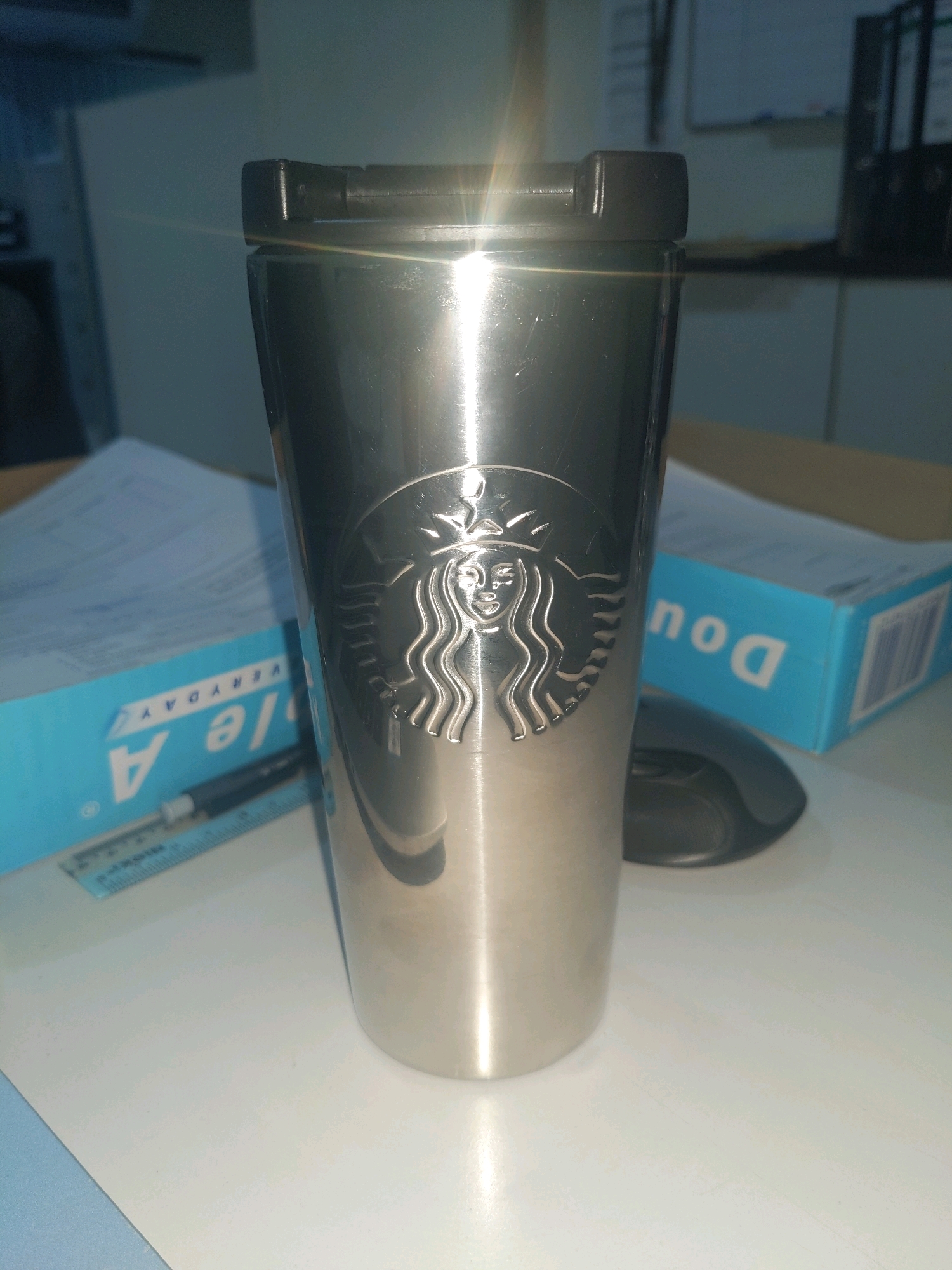 A metallic Starbucks tumbler sits upright on a white desk, reflecting bright light. The tumbler features the embossed Starbucks siren logo. Behind the tumbler, a blue box of paper with the label "A" and "EVERYDAY" is visible, along with a ruler and a pen. A black computer mouse is partially visible to the right of the tumbler. In the background, office shelving units are dimly lit. The scene appears to be an indoor workspace in Kolombong, Malaysia.