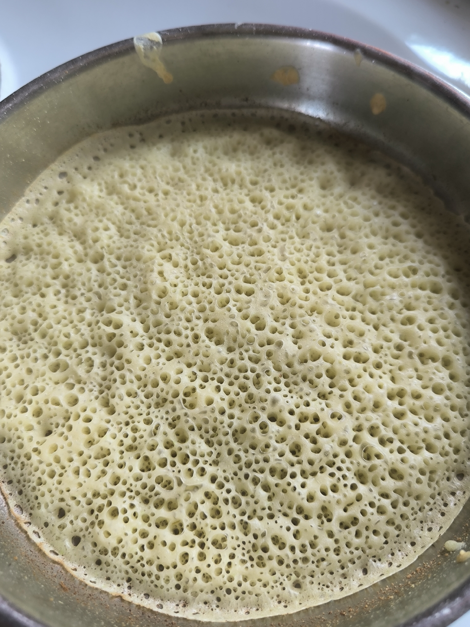A close-up, overhead shot shows a round, light-yellow pancake with a porous surface, as if a golden sponge cake has been removed from its pan. The pancake fills the bottom portion of a metallic pot, its bubbly texture filling the entire frame. The surface of the pancake is speckled with thousands of tiny circular openings, creating a textured, almost cellular pattern. A slight browning is visible around the edges of the pancake, where it meets the rim of the pot. A few small, dried droplets of batter cling to the inside of the pot's rim, appearing as yellow splotches. The background is a blurry, light-colored surface, suggesting a kitchen countertop. There are no individuals present in the image. The scene does not offer any specific indicators that would place it in Maiskii, Russia.
