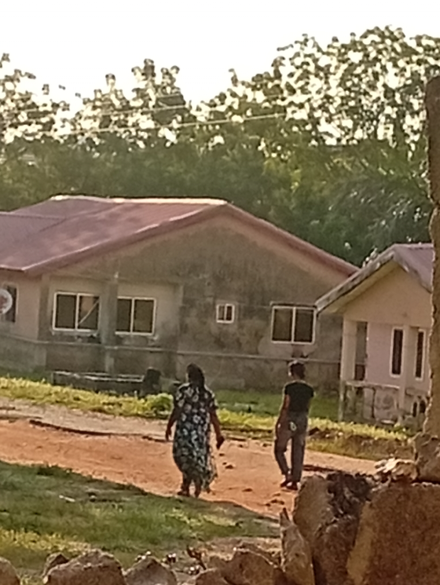 In Adankolo, Nigeria, a woman in a floral dress and a man in dark clothing walk away from the camera on a dirt path. They are in front of a weathered, single-story building with a red roof. The building has two windows visible, and another smaller, lighter-colored building is to its right. Trees are visible in the background, and the ground is covered in grass and dirt. The scene appears to be outdoors, possibly during daylight hours.