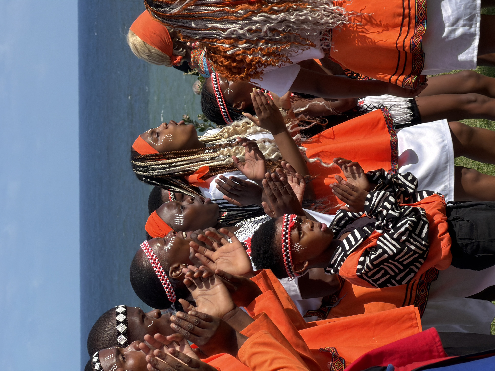 A group of people, primarily young adults, are lined up horizontally in the image, possibly performing. They are wearing traditional orange and white clothing with intricate designs and decorative beaded headbands. Some have painted white dots on their faces. They have their hands raised and are clapping or praying. Behind them, there is a body of water (possibly the ocean), with a faint line of land visible in the distance. The sky is clear and blue. The scene appears to be taken outdoors, possibly in Cape Town, South Africa, although specific landmarks are not visible to confirm this location.