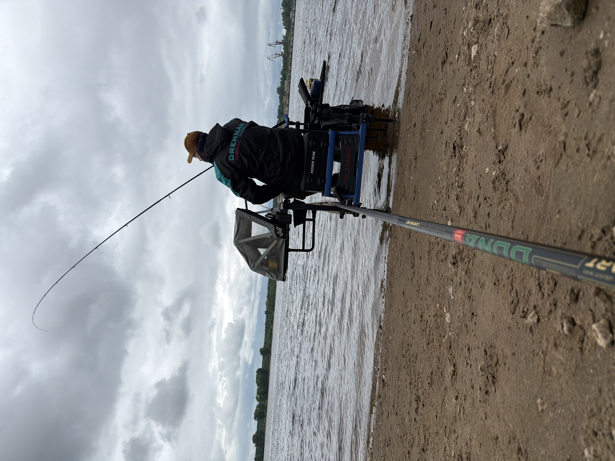 A person is engaged in feeder fishing on the sandy shoreline of a large body of water, likely a river or reservoir, under an overcast sky in Chaikovsky, Russia. The individual, viewed from the rear, wears a black and teal jacket with "DRENNAN THERMAL" printed on the back and an orange baseball cap. They are seated on a multi-legged fishing platform, or "seat box," which extends into the shallow water at the edge of the shore. The seat box features labels including "FEEDER TEAM" and "VOLZHANKA." A fishing rod extends from the platform upwards and to the right, with its tip noticeably curved, suggesting tension. Another fishing rod, marked "DUNA ART" and partially "LANDING NET," is positioned diagonally across the sandy foreground, resting on the ground. To the left of the angler, a grey fabric cover or canopy is attached to the fishing platform. The water surface exhibits ripples, and a distant treeline is visible on the opposite shore. In the far right background, two large industrial cranes are discernible, indicating a port or industrial area. The sky is uniformly grey with thick clouds, and the ground consists of damp, light brown sand with scattered small rocks.