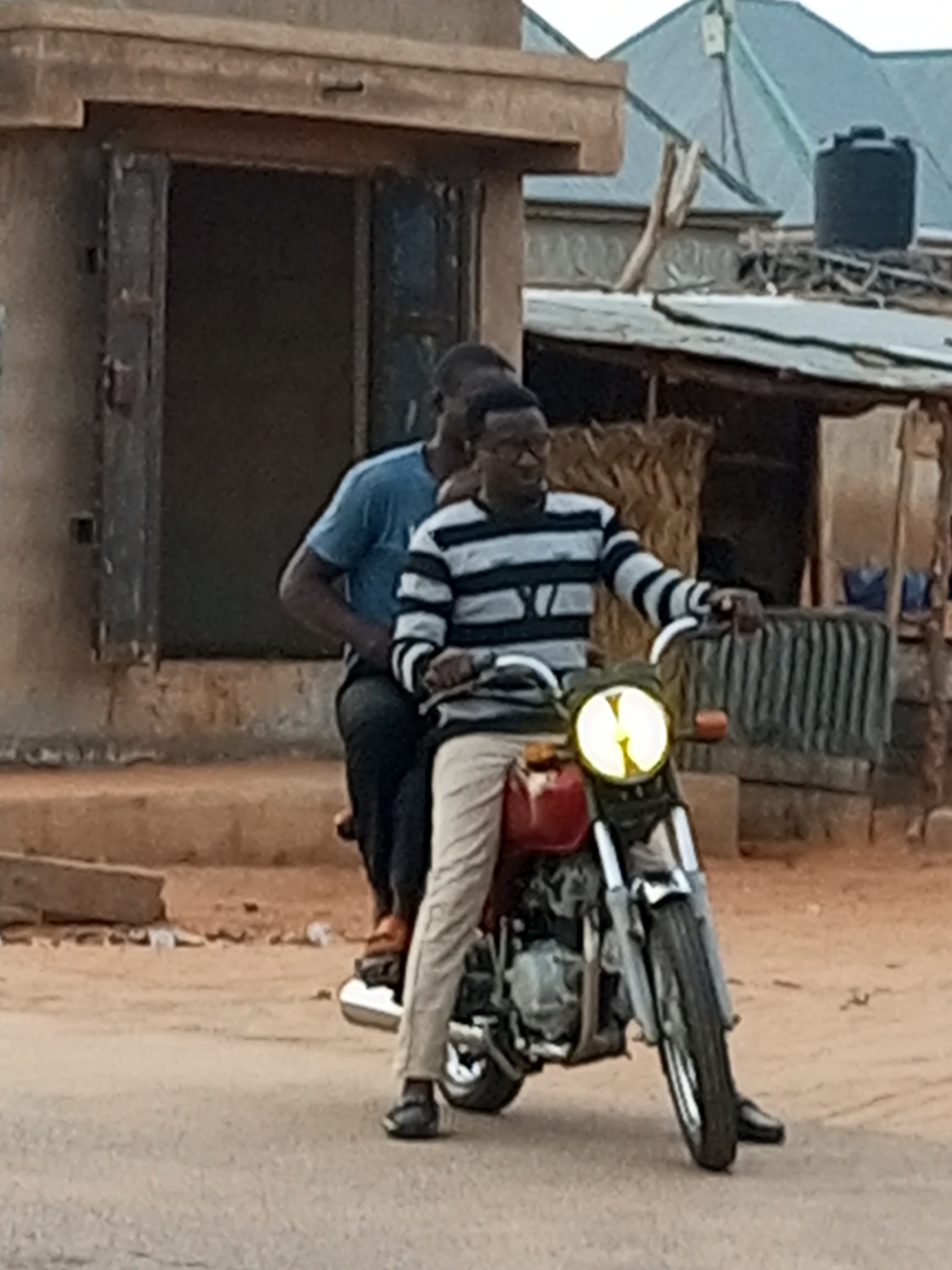 Two individuals are on a red motorcycle in what appears to be an urban or semi-urban environment in Tempure, Nigeria. The person in the front, who is driving, is wearing a black and white horizontally striped long-sleeved shirt and light-colored pants. They have glasses and their hands are on the handlebars. The person in the back is wearing a blue t-shirt and dark pants. They are seated behind the driver. The motorcycle has a prominent circular headlight that is illuminated. The background consists of a building with a wooden door, a corrugated metal roof structure, and some vegetation. The ground is a sandy, reddish-brown dirt road.