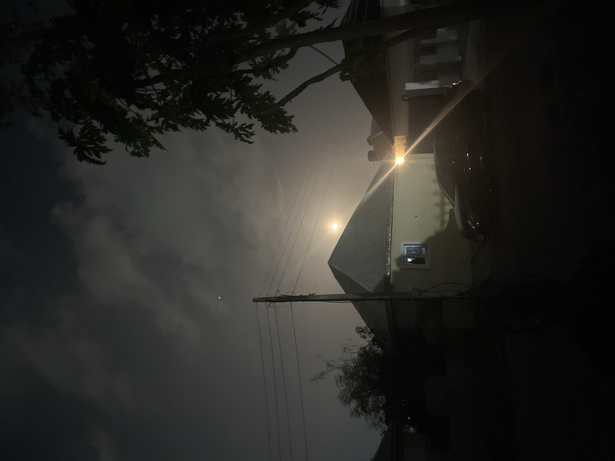 The image, taken at night, shows a residential scene in Abuja, Nigeria. A two-story building is visible on the right, lit by a street light above. Power lines run across the foreground, intersecting the faint moon visible through the cloudy sky. Silhouetted trees partially obscure the view on the left. There are no visible people or actions. The environment appears to be a typical urban neighborhood in Abuja.