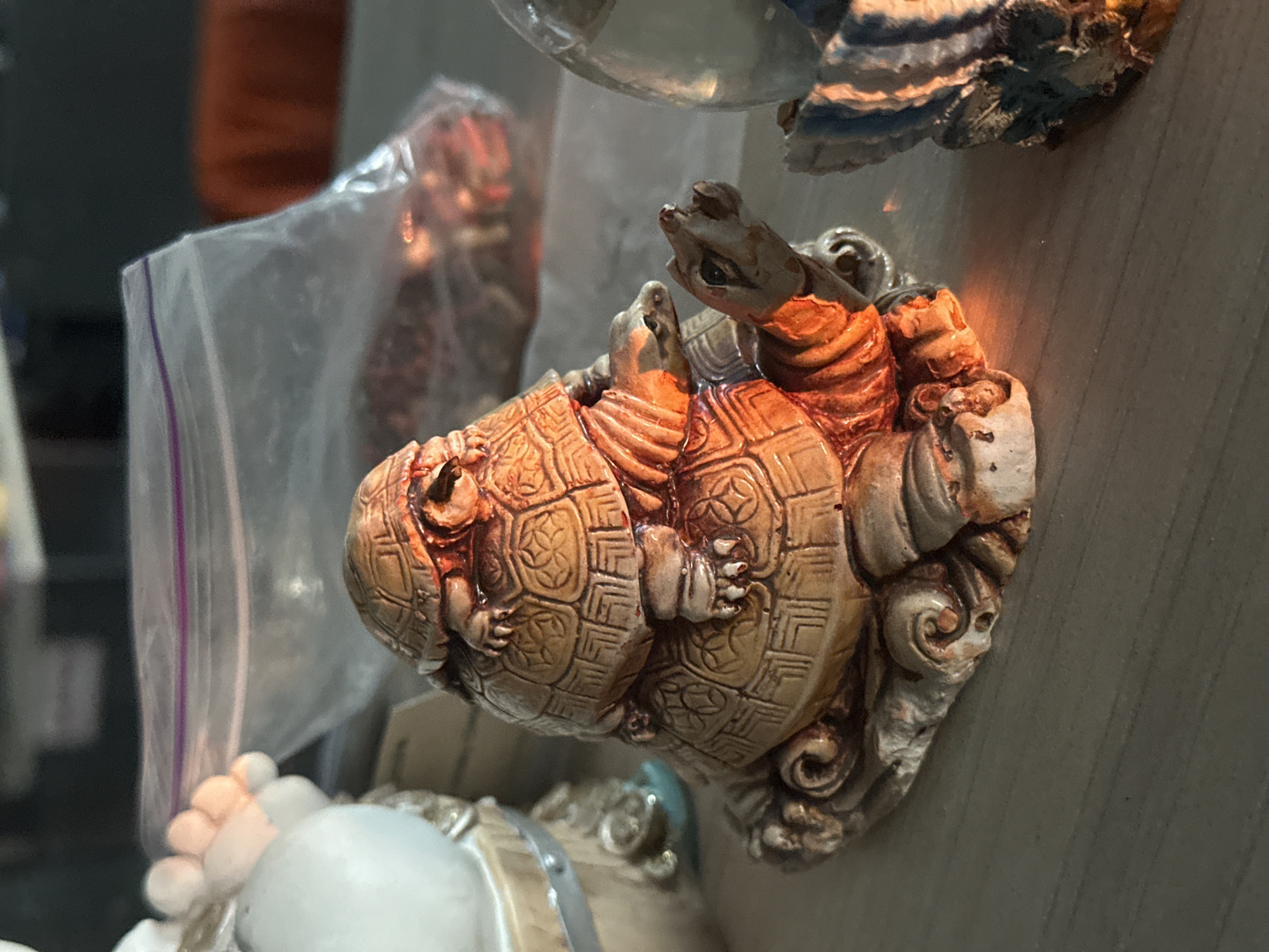 The image is a close-up, medium shot of a stack of turtle figurines. The turtles are a light brown color with darker brown detailing on their shells and limbs, and they appear to be made of ceramic or resin. The largest turtle is at the bottom, followed by a medium-sized turtle, and then a smaller turtle on top, giving the appearance of a pile. The turtles are stacked on a textured base that resembles rock or sand.
To the left of the turtle figurines, a portion of another figurine is visible. It appears to be a light-colored, possibly white, object with several rounded protrusions, resembling toes or fingers. Behind the turtles, a clear plastic bag with a purple zipper closure is visible. Inside the bag, there appear to be small, dark objects. Further in the background, a blurry orange surface is visible. To the right of the turtles, another figurine is partially visible, colored in shades of blue and gray with a textured surface.
The figurines are placed on a flat, light brown surface that appears to be a wooden table or shelf with visible grain. The lighting in the image is warm and casting shadows, with a bright orange glow illuminating the lower right side of the turtle figurines, suggesting a light source is present. The overall scene is indoors, with a shallow depth of field blurring the background elements.
The location in the city of Hora, Ukraine is not discernible from the provided image.