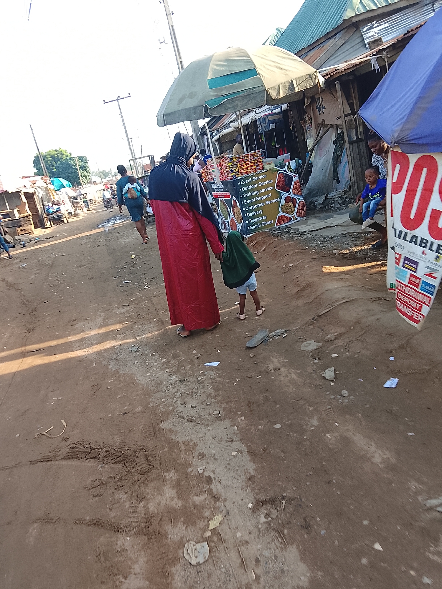 The image portrays a street scene in Abuja, Nigeria. The unpaved road is lined with informal shops, indicated by their makeshift structures and signage. A woman in a black hijab and red gown holds the hand of a child dressed in green and light-colored shorts, walking away from the camera. A poster with red letters and white text is visible on the right side of the frame. The poster indicates "Withdrawal Deposit Transfer." Additional people and vehicles are visible further down the road, suggesting a busy market area.