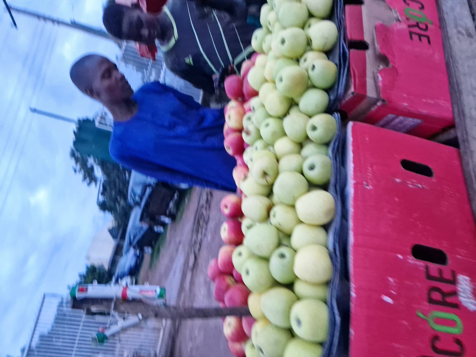 The image depicts an outdoor fruit stall in the city of Kabong, Nigeria, likely during daylight hours given the ambient lighting. A prominent display of fresh apples, both red and green varieties, is arranged in crates. Two young males are visible near the apples; one, wearing a blue traditional garment, appears to be looking down towards the fruit, while the other, in a dark t-shirt with a striped design, is partially obscured. In the background, various vehicles are parked, some covered with tarps, and a metal fence is also visible. The overall scene suggests a commercial setting, possibly a market or roadside vendor.
