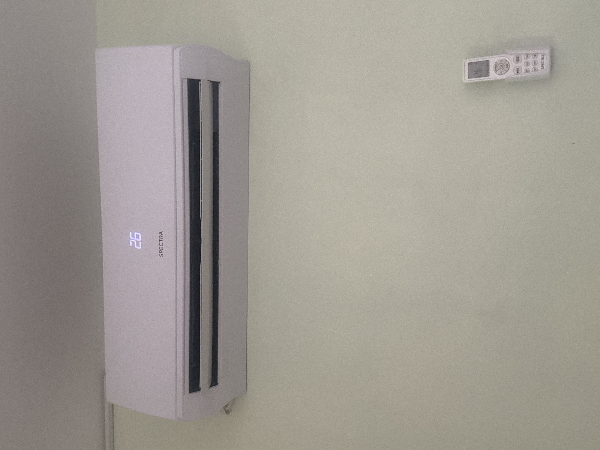 An air conditioning unit, branded "SPECTRA," is mounted on a light green wall. The unit displays a temperature of "26". A white remote control, also branded "SPECTRA," is shown on the wall to the right of the air conditioning unit. The remote control also displays a temperature of "26". The scene appears to be indoors, likely a residential or commercial space. No people are present in the image. The location is not specified as Lod, Israel in the image itself.