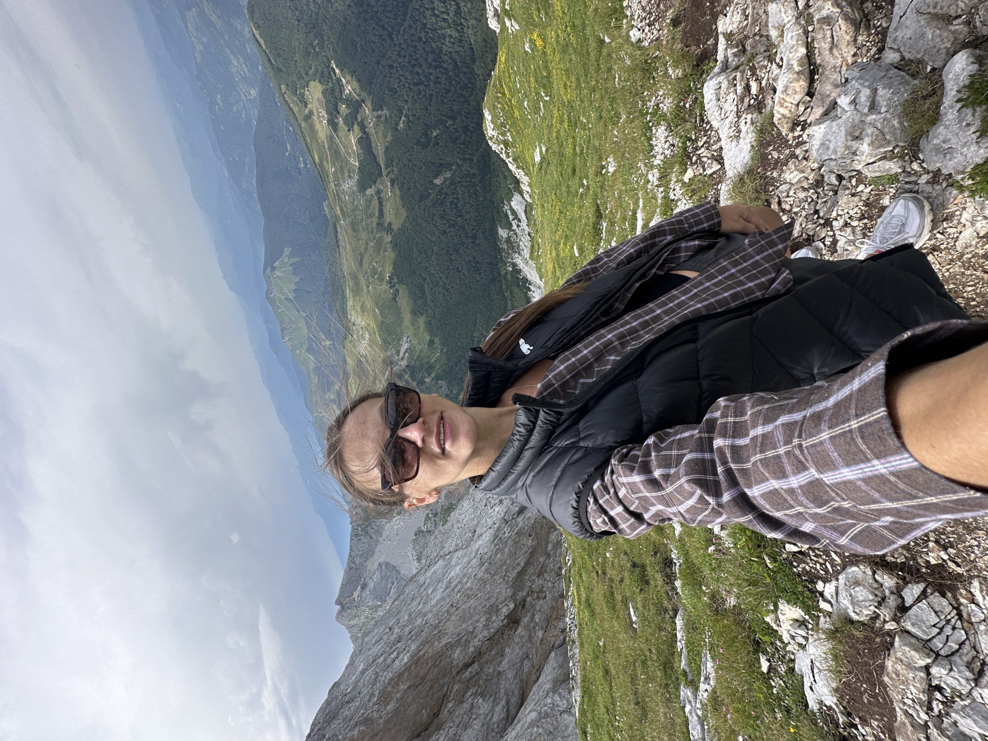 In the image, a woman with brown hair and sunglasses is taking a selfie while standing on a rocky, grassy mountainside. She is wearing a black puffer vest over a brown and white plaid shirt. Her right arm is extended, holding the camera. Behind her, a vast, mountainous landscape unfolds, with layers of green hills and distant, hazy mountains under a cloudy sky. The terrain is rugged, with large rocks and patches of grass. The specific location in the city of Unknown, Montenegro, is not discernible from the visual information.