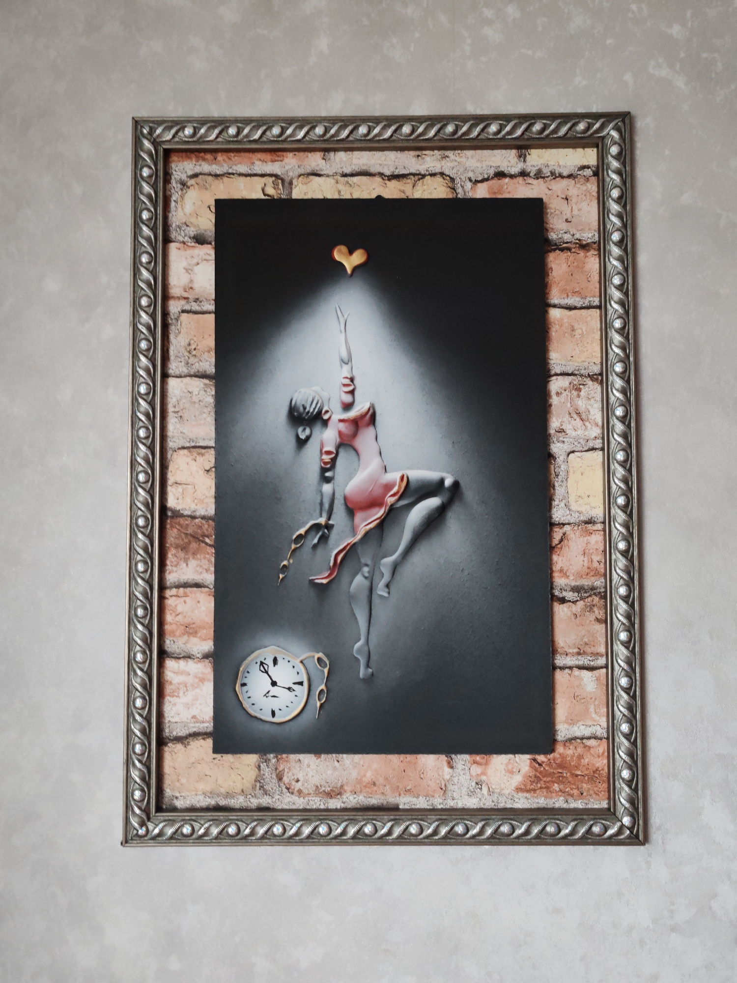 The image shows a framed artwork hanging on a wall. The artwork depicts a surrealist scene with a figure that appears to be a dancer, rendered in a sculpted or bas-relief style. The dancer is wearing a pink dress, and a golden heart-shaped object is positioned above her head. A melting clock, reminiscent of Salvador Dali's work, is located at the bottom left of the artwork. The artwork is set against a gray backdrop and appears to be illuminated by a spotlight.
The frame surrounding the artwork is silver and ornate. A brick-patterned material is visible between the artwork and the frame, suggesting a layered or decorative effect. The wall on which the artwork is hanging has a textured, neutral-toned finish.
There is no information to suggest that this image was taken in Petropavlovsk-Kamchatsky, Russia.
