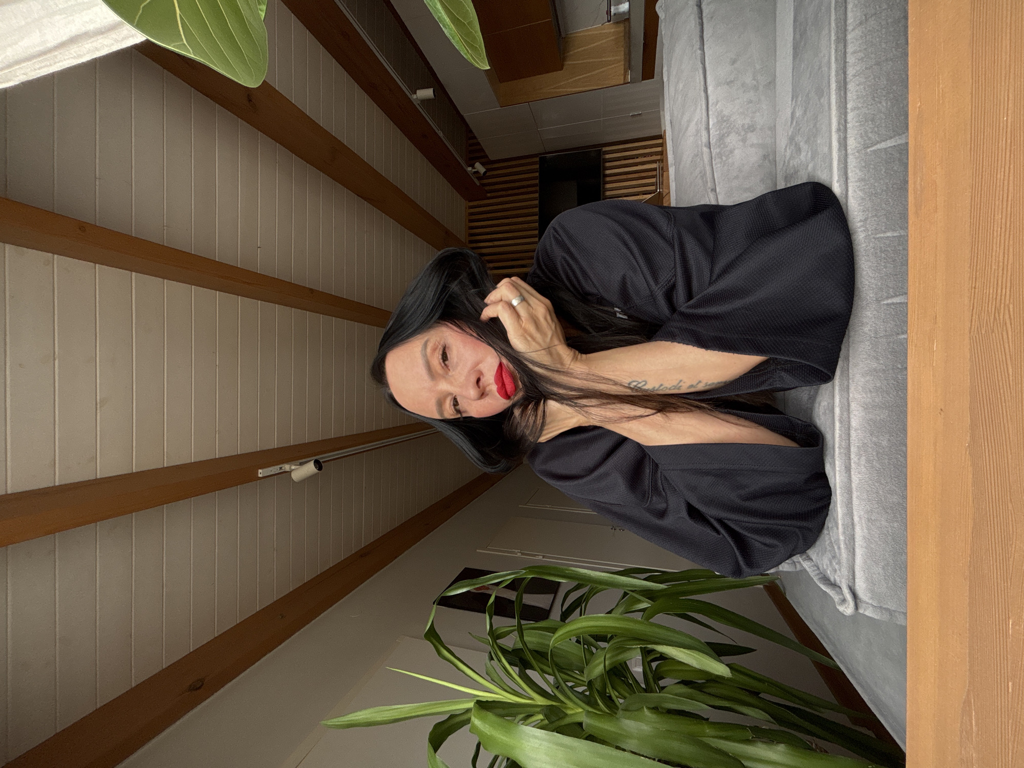The image captures a woman posing indoors in a residential setting, likely in Chaikovsky, Russia. The woman, with long dark hair and red lipstick, wears a black textured top and a silver ring on her left ring finger. She has a tattoo on her left forearm that reads "Custodi et". She is looking directly at the camera with a relaxed expression, her left hand near her face, possibly touching her hair.
She is leaning against a light gray, upholstered surface, which could be a sofa or a padded wall. In the foreground, a large houseplant with long, green leaves adds a natural element to the scene.
The room features distinctive architecture, including a sloped ceiling with prominent wooden beams and white vertical paneled walls. A modern light fixture with a silver bar is mounted on the upper wall paneling. In the background, another part of the interior is visible with darker wooden slat walls, a dark rectangular object (possibly a screen), and a white toilet roll dispenser. The lighting appears soft and diffused, indicating daytime. The overall impression is one of a comfortable and styled interior.