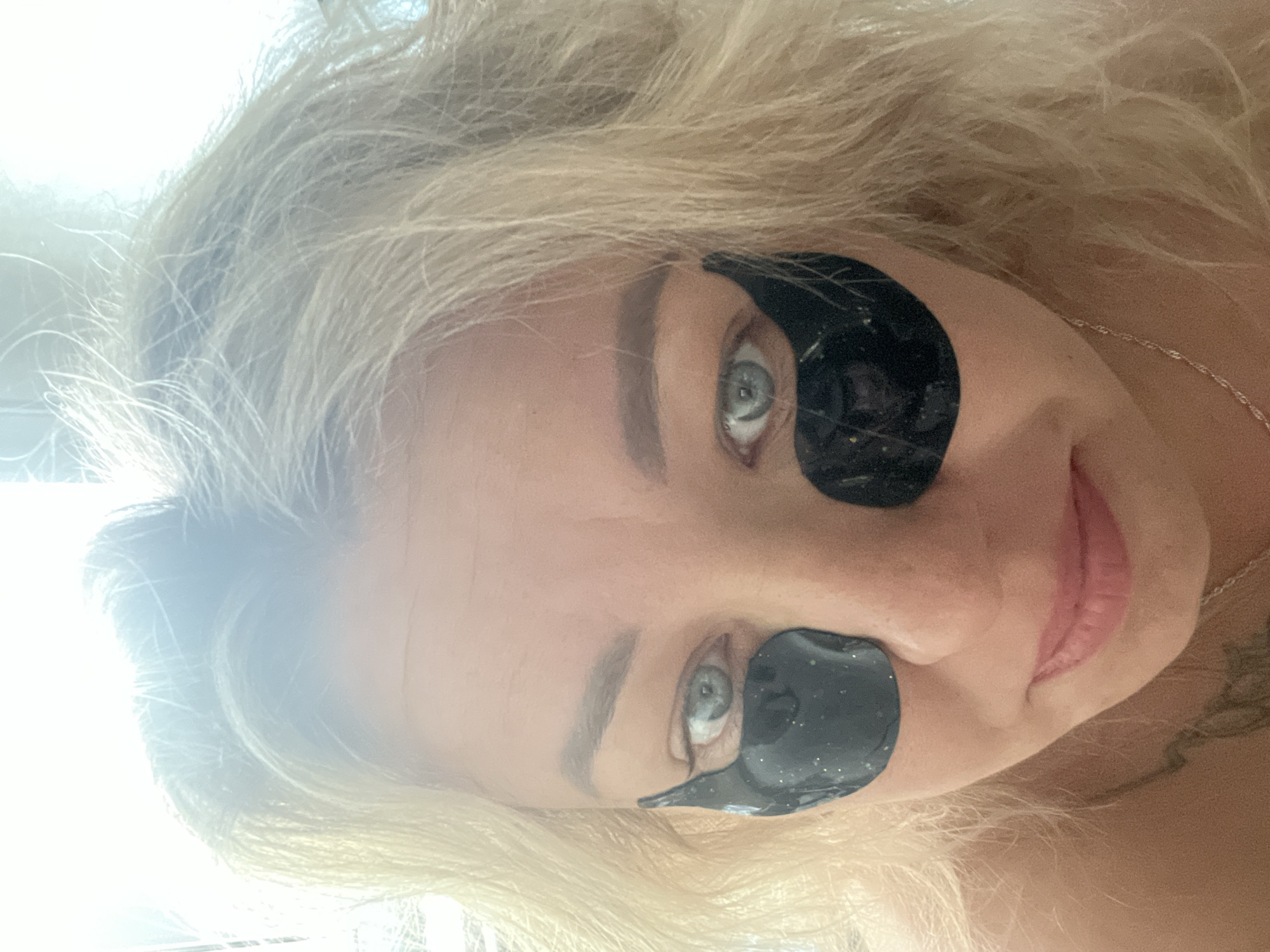 A woman with blonde hair and blue eyes is pictured from a close, rotated angle. She has black eye masks under each eye. A silver necklace is visible. Part of a tattoo is visible on her neck. The location of Chaikovsky, Russia is not discernible from the provided image.
