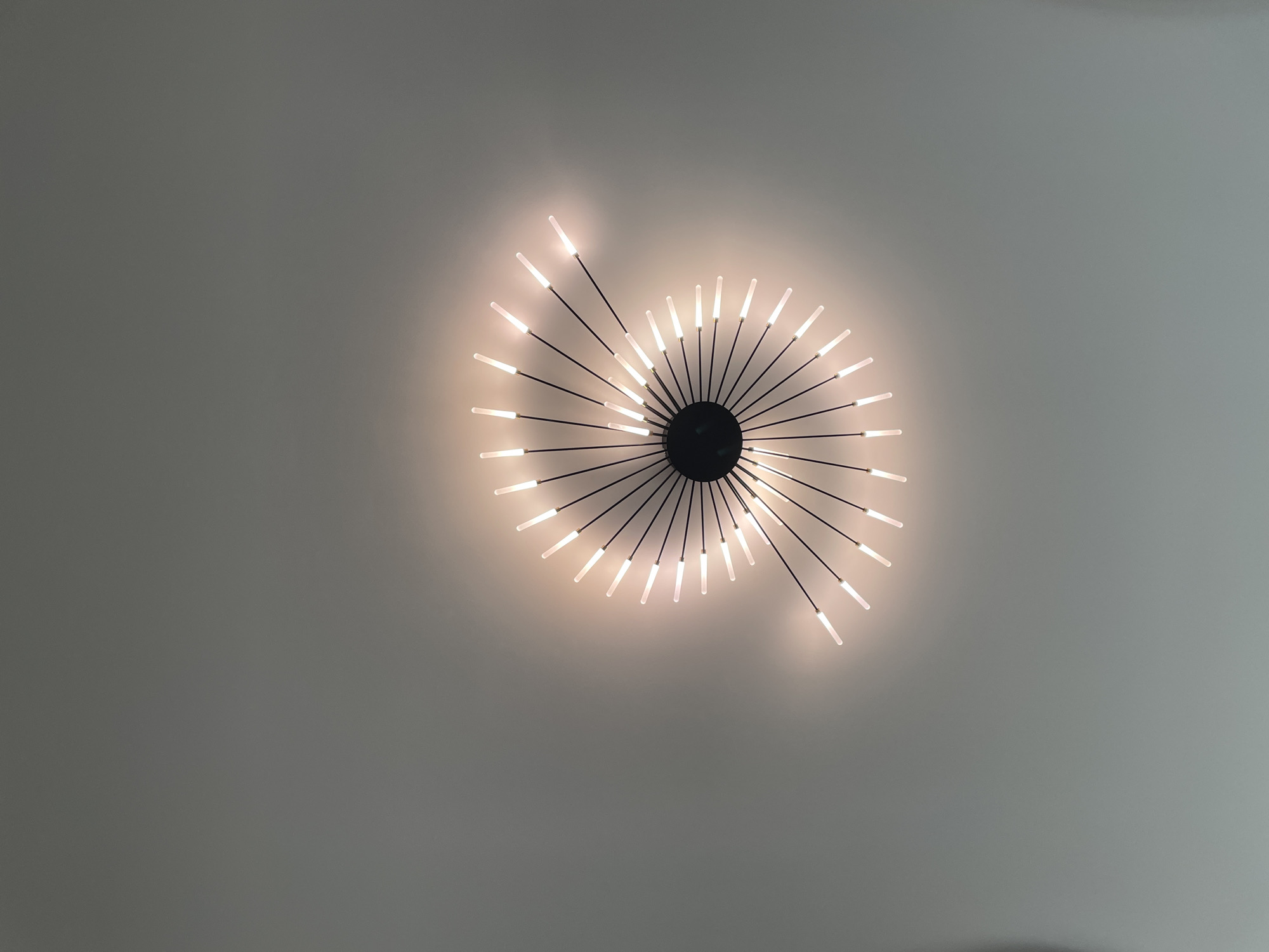 A light fixture is affixed to a ceiling. The fixture consists of a dark, circular center from which multiple rods radiate outward. Each rod ends in a cylindrical light, producing a starburst pattern against the light grey ceiling. The image is taken from below, looking up at the fixture. The scene is located in Лесной Пейзаж Кп (шарапово), Russia.