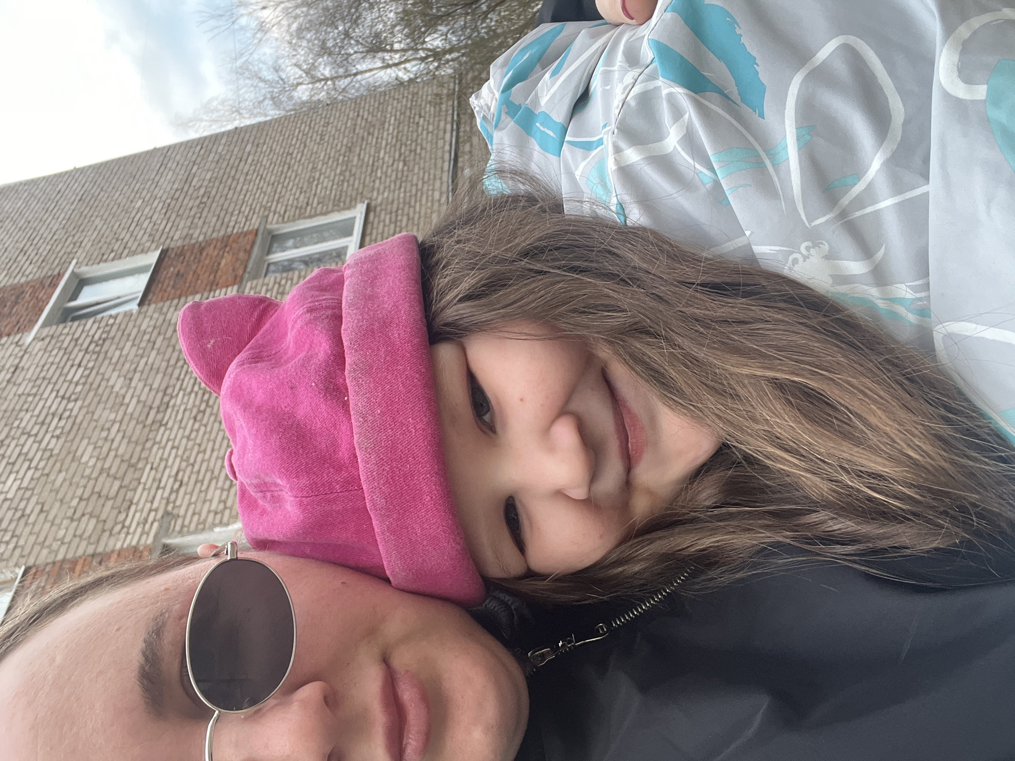 This is a close-up, tilted selfie taken outdoors in Chaikovsky, Russia, featuring two individuals under an overcast sky.
In the foreground, on the right side of the frame, is a young child with long, light brown hair, wearing a vibrant pink hat or hooded garment. The child is smiling broadly and looking directly at the camera, conveying a happy and content expression. Their head appears to be resting on the shoulder or chest of the second individual.
The second individual, likely an adult, is positioned on the left side of the frame, with only a portion of their face visible. They have light brown hair and round, dark-lensed sunglasses are pushed up onto their forehead. Their lips are slightly parted, and their expression is neutral. They are wearing a dark jacket, possibly black, with a silver zipper visible. Another garment with a light grey background and abstract light blue and white patterns is partially visible in the upper right.
The background features a large, light-colored brick building with several rectangular windows, suggesting an urban or residential setting. Above the building, bare tree branches are visible against a bright, cloudy sky, indicating a cooler season like winter or early spring. The image captures a casual, intimate moment between the two subjects. No visible text is present in the image.