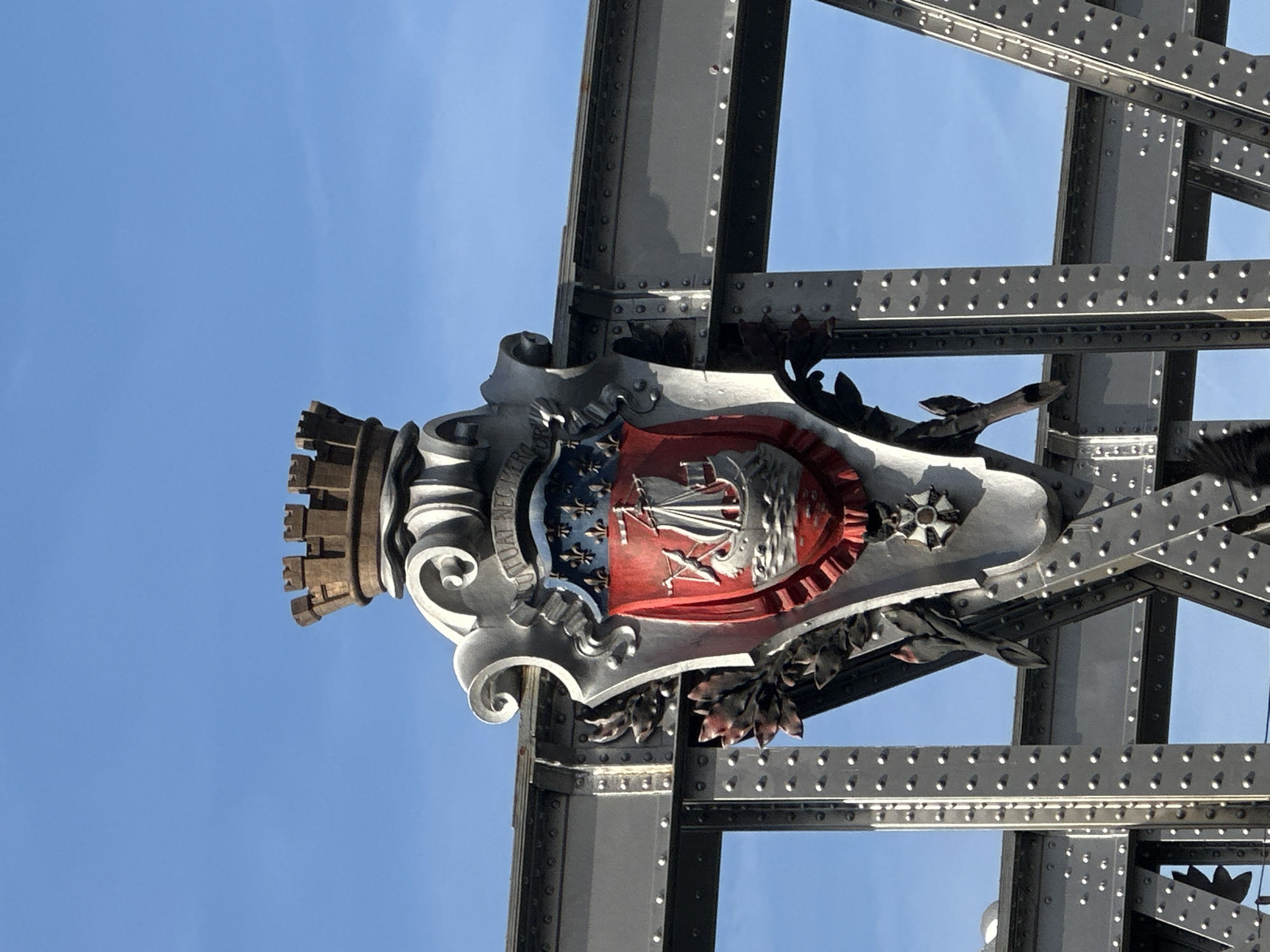 The image shows a close-up, eye-level, medium shot of a decorative crest mounted on a gray metal bridge structure, set against a clear blue sky. The crest features a golden crown atop a white, ornate shield. The shield itself is divided into sections, with the upper part displaying a blue background adorned with gold fleur-de-lis symbols. The lower, larger section of the shield is red and depicts a white sailing ship with three masts and furled sails, navigating choppy gray waves. Two dark green laurel branches are attached to the sides of the shield, and a small white and black star-shaped emblem is visible at the bottom. The bridge structure is made of dark gray metal beams and is fastened with numerous rivets. The crest is likely a coat of arms, possibly representing a historical or civic entity associated with Paris, France. The overall scene is static, with no people or activity observed.