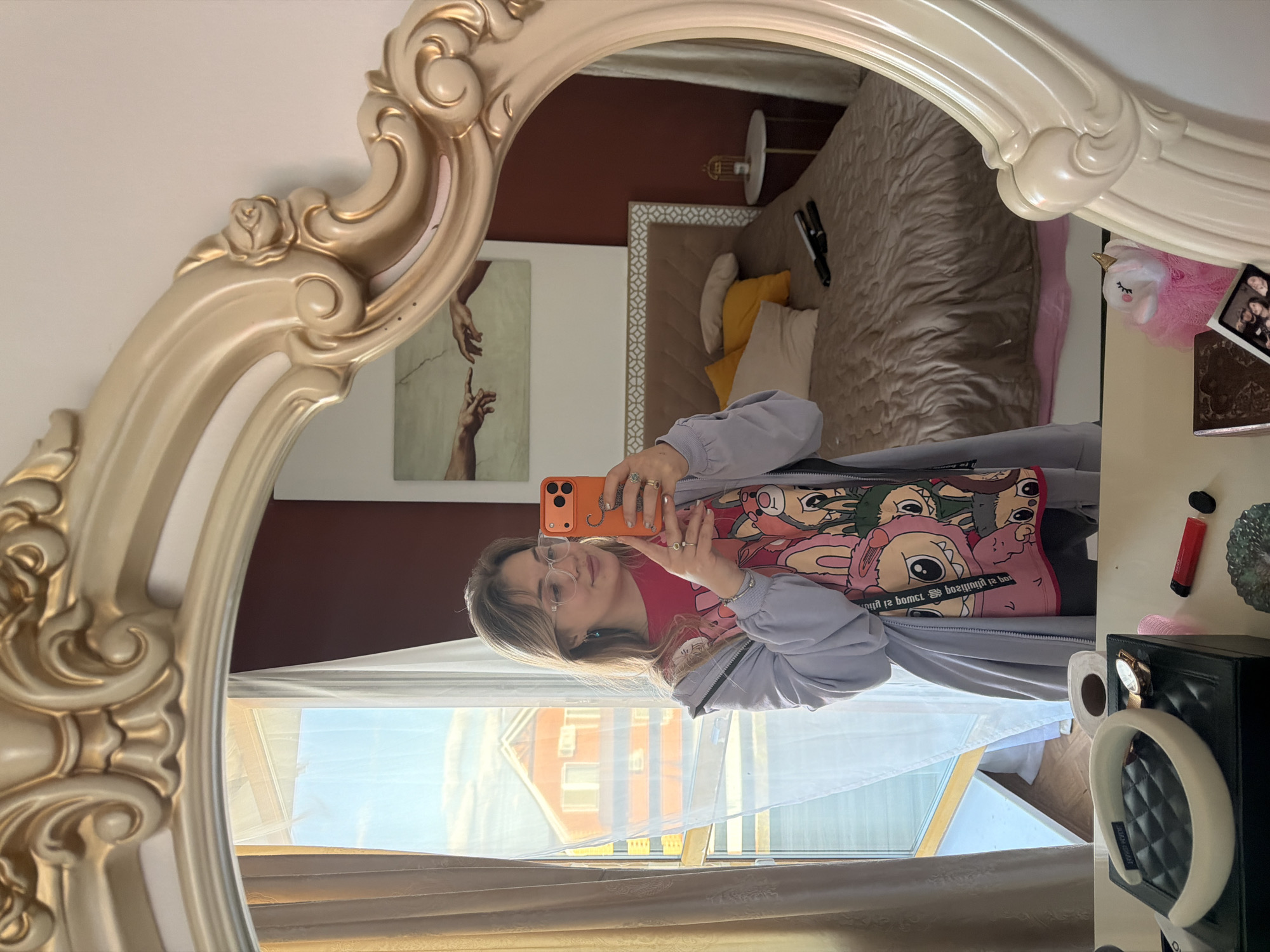 A person is seen taking a mirror selfie in what appears to be a bedroom. The image is taken from a low angle, reflecting the person and elements of the room. The mirror has an ornate, cream-colored frame with gold accents and decorative carvings including a rose design.
The subject is a young person, likely female, with light brown hair, wearing glasses, a grey zip-up jacket or hoodie over a red t-shirt featuring a colorful cartoon graphic with large-eyed characters, possibly from an animated series. They are holding an orange smartphone with an orange case, displaying rings on their fingers.
The reflection shows the room behind the person. To the left, a painting resembling "The Creation of Adam" with two hands reaching out is visible on a light wall. Further back is a bed with a beige quilted bedspread and a headboard with a geometric pattern, adorned with yellow and white pillows. A small, golden-based lamp with a white shade stands beside the bed. The wall behind the bed is a dark red or burgundy color.
To the right, a white dresser or vanity is partially visible, holding various items: a black quilted jewelry box or case, a white headband, a wristwatch, a roll of toilet paper, a pink fluffy unicorn toy, a small photo frame or tablet, and a red lip gloss tube.
Through a window covered by sheer white curtains, an outdoor scene is visible. A multi-story building with orange-red brick walls and lighter colored windows can be seen under a bright blue sky, indicating daytime. Some text is visible on the person's t-shirt, although mirrored and partially obscured, and some faint text might be on the black box, but it is unreadable.