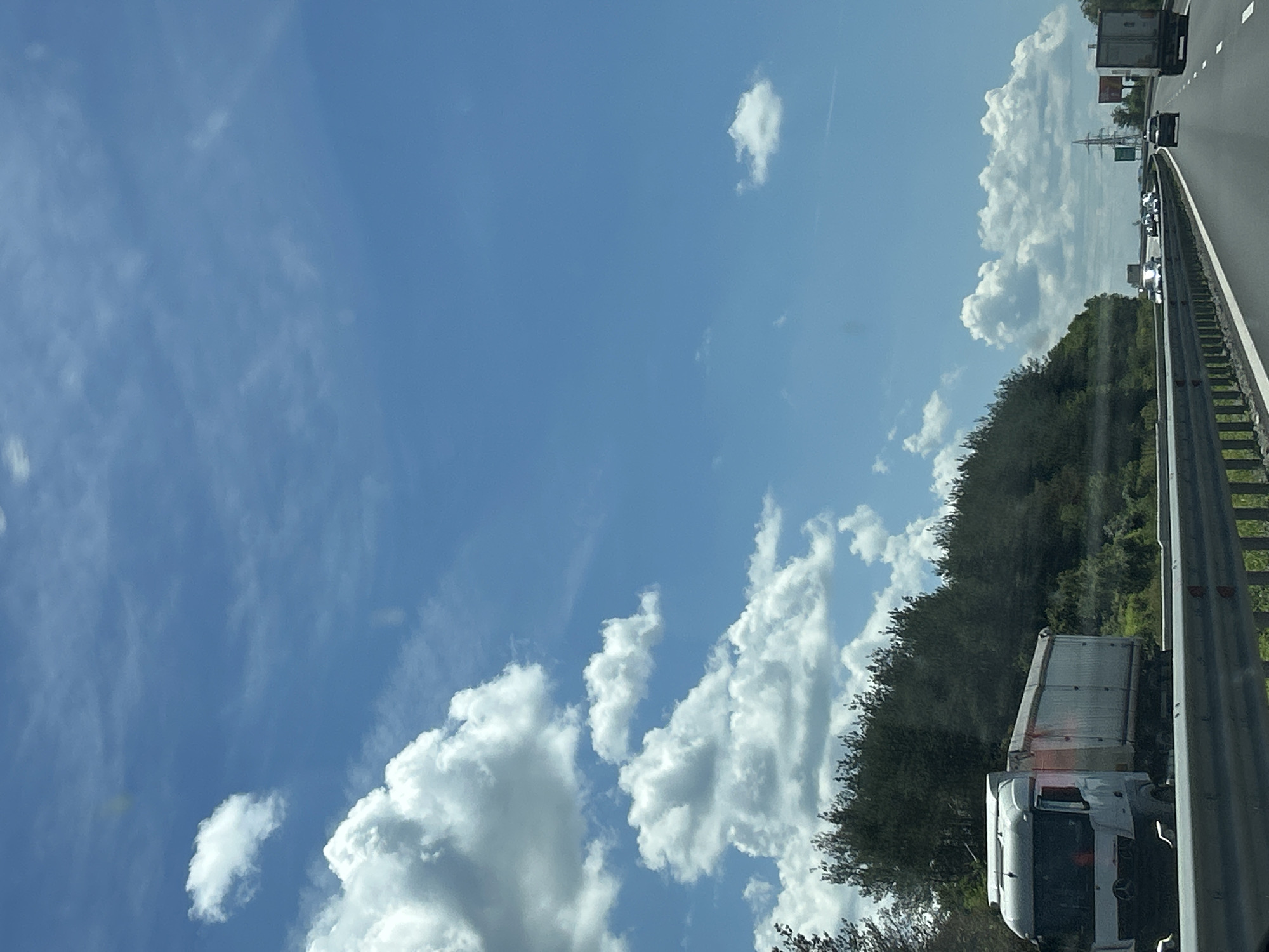 The image depicts a highway scene on a sunny day with scattered clouds in a bright blue sky. A white truck with a silver trailer is in the foreground on the left side of the highway, traveling in the same direction as the camera. The truck is on the left lane, and to its right, there are other vehicles on the highway. A dense line of green trees and vegetation runs along the left side of the highway, forming a hilly landscape. In the distance, on the right side of the highway, there are power transmission towers and signs. The highway has multiple lanes, and a metal guardrail is visible in the lower part of the image. The overall impression is a typical highway journey through a natural landscape. The location is identified as the highway near Rajana, Bulgaria.