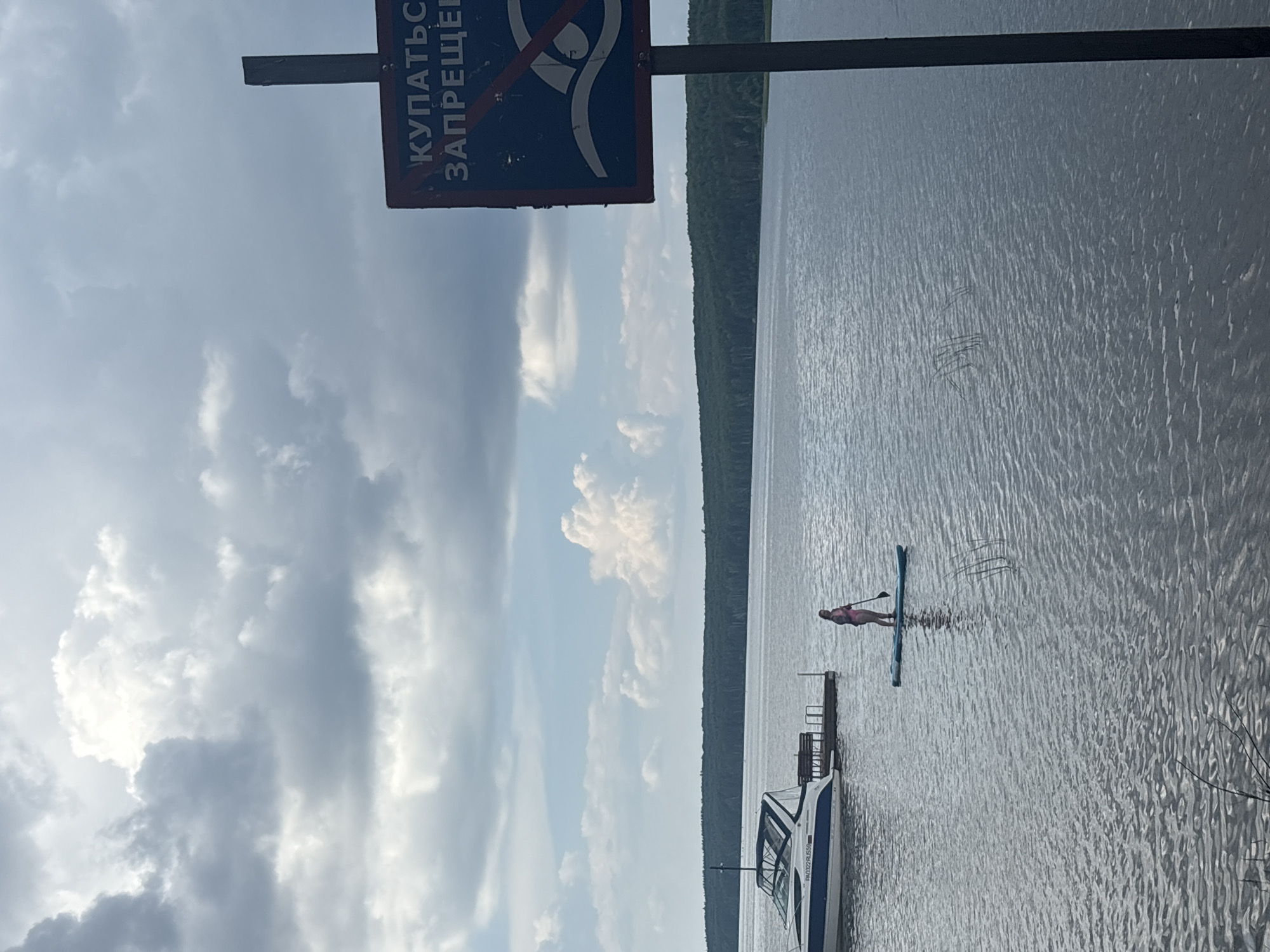 A person is paddleboarding on a large body of water with a boat docked nearby and a forest in the background. A sign in the foreground indicates that swimming is prohibited. The sky is cloudy.