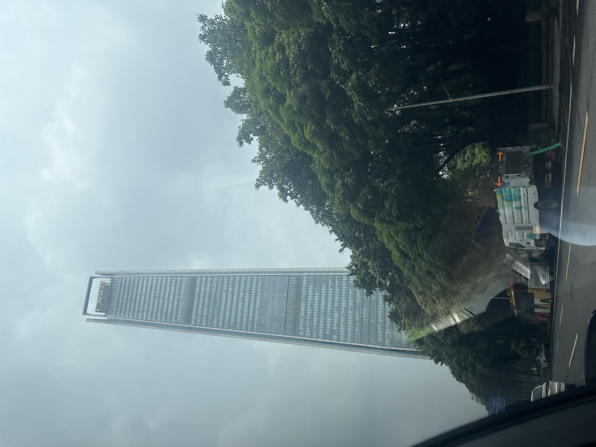 The image captures a street scene in Tianhe District, China, on an overcast day. A towering, modern skyscraper with a glass facade dominates the background, its top section bearing golden Chinese characters. Lush green trees line the right side of the street, obscuring some of the lower levels of the building. In the foreground, a road with white and yellow lane markings is visible. Several vehicles are present: a white car is partially visible on the left, a bus with red, yellow, and white sections is in the middle, and a white street cleaning truck with a green hose extending to the ground is to the right of the bus. The truck has colorful circular patterns and an orange arrow display on its rear. The overall environment appears to be urban, with signs of city infrastructure like streetlights and a road surface that looks damp, suggesting recent rain or cleaning.
