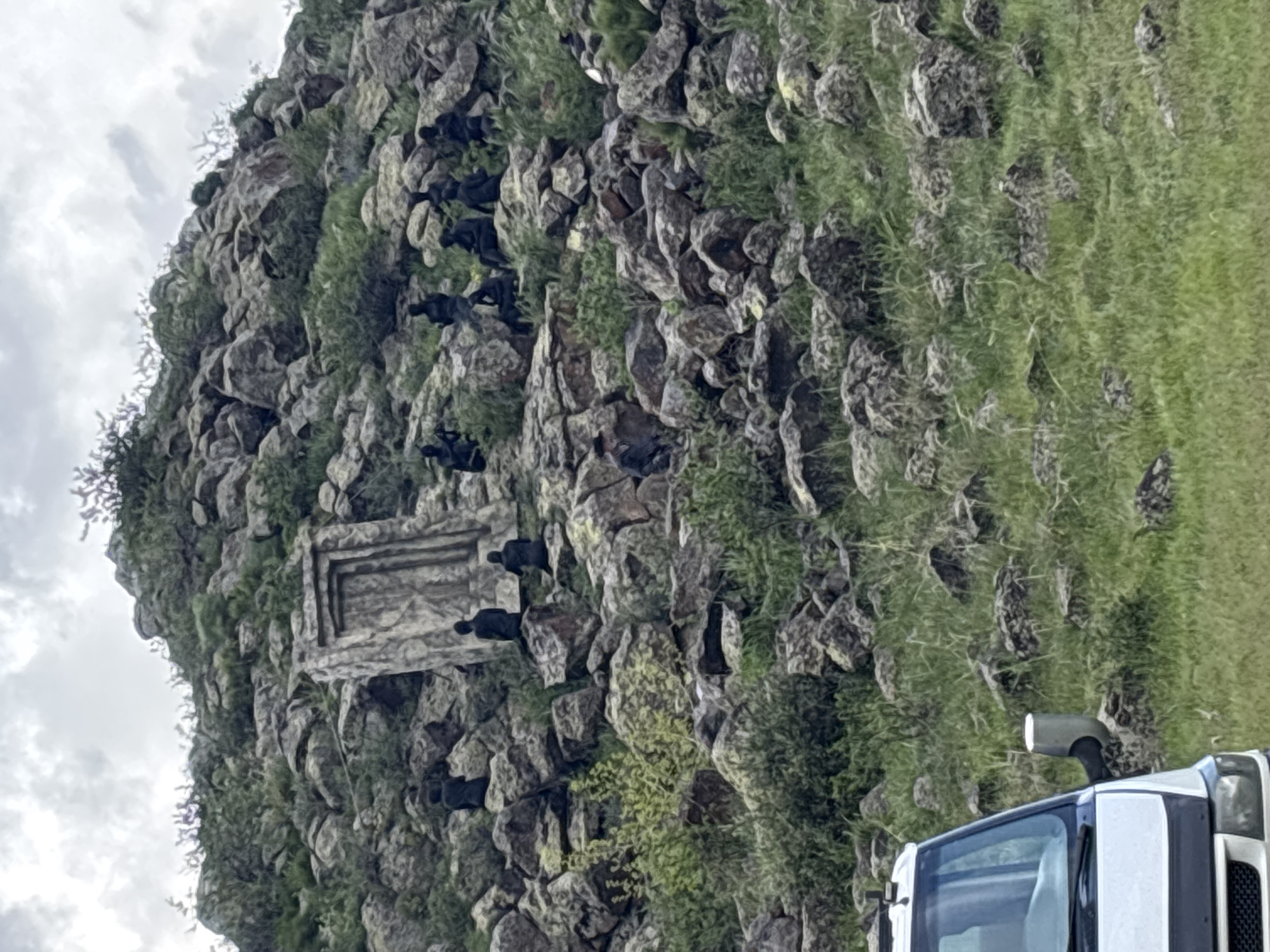 The image shows a rocky hillside in Tlik, Armenia. The hillside is covered with grass and rocks, and several black objects are scattered across the rocks, possibly animals or figures. A stone structure, resembling a doorway or frame, is embedded in the rocks on the hillside. The top of a white vehicle is visible in the lower right corner of the image. The sky is overcast.