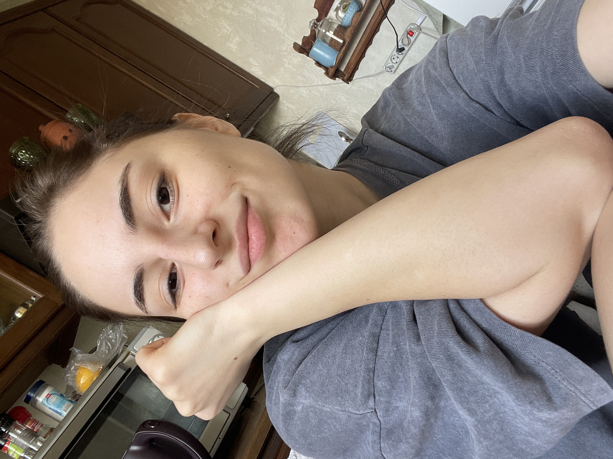 A woman with fair skin and dark hair, adorned with hair rollers, rests her face on her hand, appearing contemplative. She wears a grey t-shirt. The setting includes a kitchen environment with visible cabinets, food items, and kitchen appliances. The location in Moscow, Russia cannot be determined from the image.