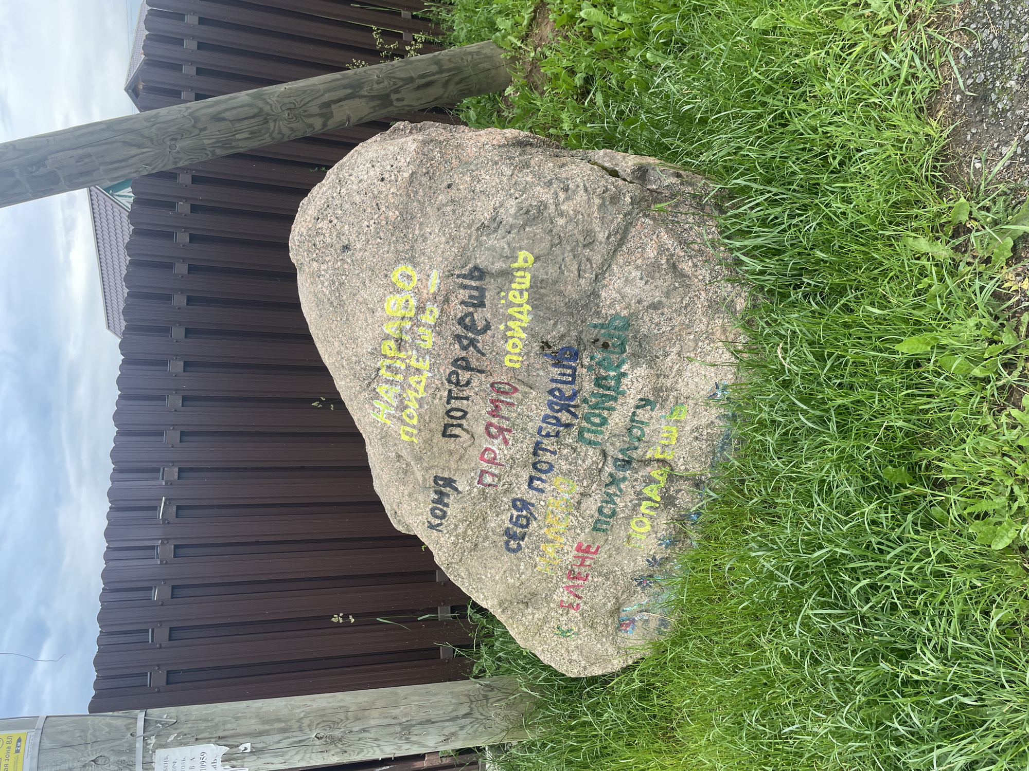 The image depicts a large, grey boulder covered in multi-colored graffiti, situated in a grassy area next to a brown, metal fence and a wooden utility pole. The graffiti on the boulder appears to be written in Russian. In the background, two houses with tiled roofs are visible. The sky is overcast. The scene is outdoors, with the boulder being the central subject, adorned with handwritten text and symbols. The surrounding environment consists of grass, a fence, and a utility pole, suggesting a semi-rural or residential setting. There are no people visible in the image.