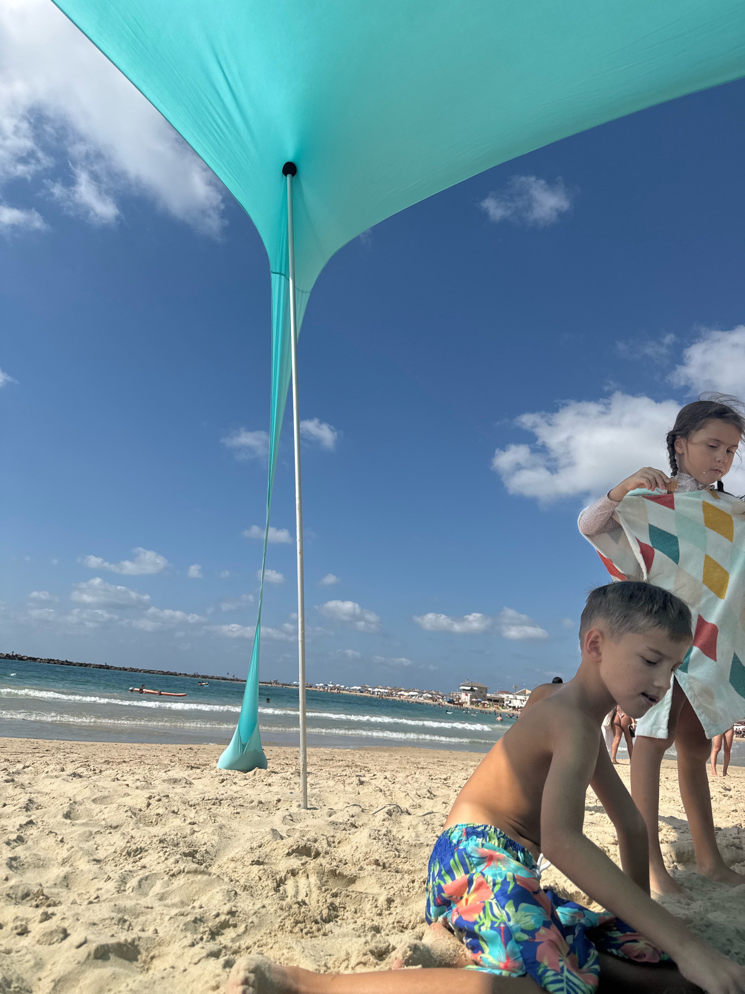 A boy with sandy hands wearing brightly colored swim trunks kneels on the sand in the foreground, looking down. Behind him, a girl with braided hair stands holding a towel with a diamond pattern. They are on a sandy beach in Netanya, Israel, with the blue sea and a breakwater visible in the background. A large, turquoise beach tent with a white pole casts a shadow over the scene. The sky is blue with scattered white clouds.