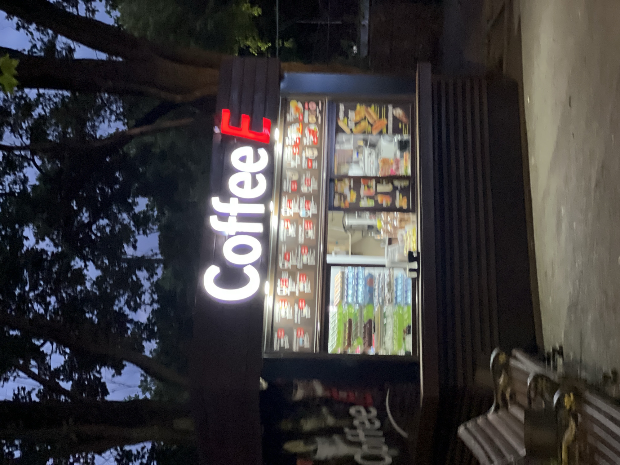 The image shows a coffee kiosk at night in the city of Krasnodar, Russia. The kiosk is dark brown with a neon sign that reads "Coffee E" in white and red letters. Inside the kiosk, there are displays of food and drinks. To the left of the kiosk, there is a bench. The background consists of trees and a dark sky.