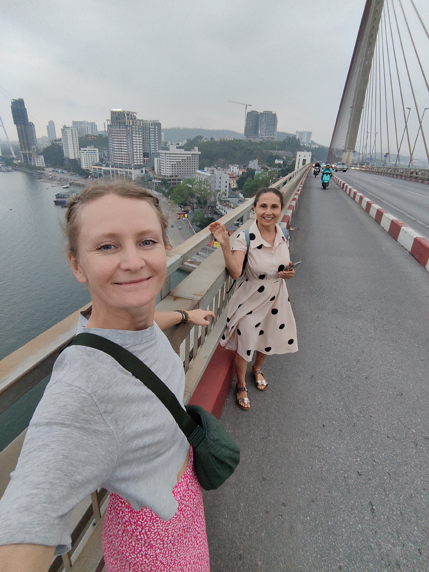 Two women stand on Bai Chay Bridge in Bai Chay Ward, Vietnam, posing for a selfie. The woman in the foreground wears a gray t-shirt, a pink skirt, and a green shoulder bag. The woman in the background wears a beige dress with black polka dots and brown sandals. She is holding a cell phone and waving. The bridge has a red and white barrier, and traffic moves along the roadway. Buildings can be seen in the background along the coastline. The sky is overcast.