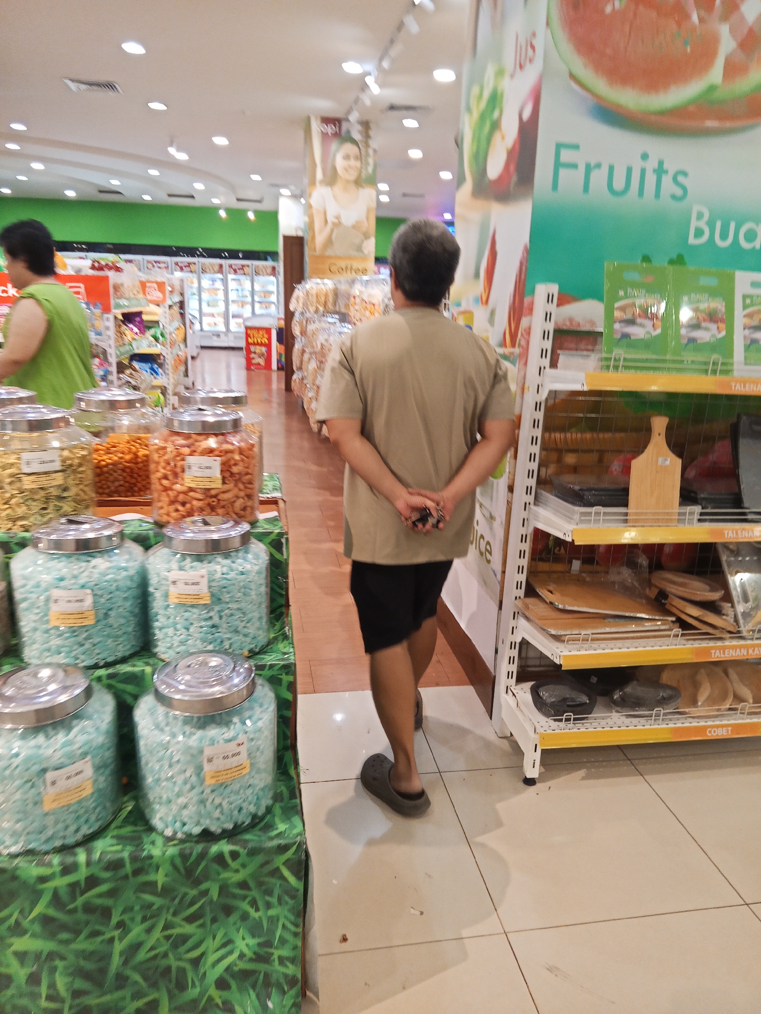 The image depicts the interior of a supermarket, likely a section selling snacks and household items. In the foreground, a person wearing a light brown t-shirt and black shorts is walking away from the camera, with their hands clasped behind their back. To the left, several large glass jars filled with colorful candies or snacks are displayed on a table covered with a green, grass-patterned cloth. Behind these jars, shelves stocked with various items are visible. To the right, a white shelf unit with yellow trim displays kitchenware, including cutting boards and what appear to be placemats or small plates. The background reveals more of the supermarket, with aisle displays, illuminated signs, and a large poster featuring fruit imagery. The overall environment suggests a well-lit commercial space. The image is not specific to Lubuklinggau, Indonesia, as there are no identifying landmarks or contextual clues to confirm this location.