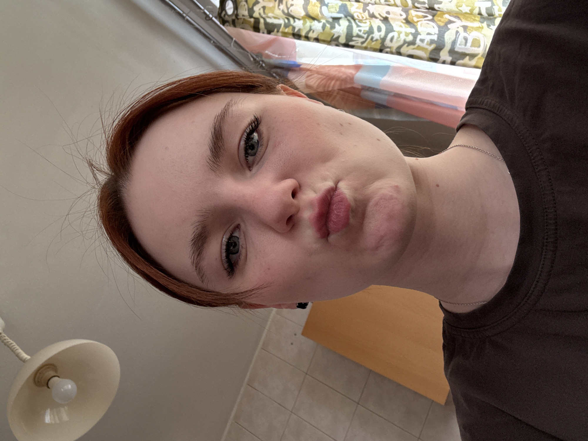 A selfie taken at a slightly upward angle depicts a fair-skinned individual with reddish-brown hair and green eyes. The individual is wearing a dark brown t-shirt and a delicate silver chain necklace. The person's lips are pursed.
Behind the individual, the scene is indoor. There is what appears to be a yellow-tinted desk or countertop, tiled flooring, and a wall. An overhead light fixture is visible in the lower left corner. In the upper portion of the image, a window with patterned curtains can be seen.
The location appears to be a private residence in the city of Unknown, Unknown.