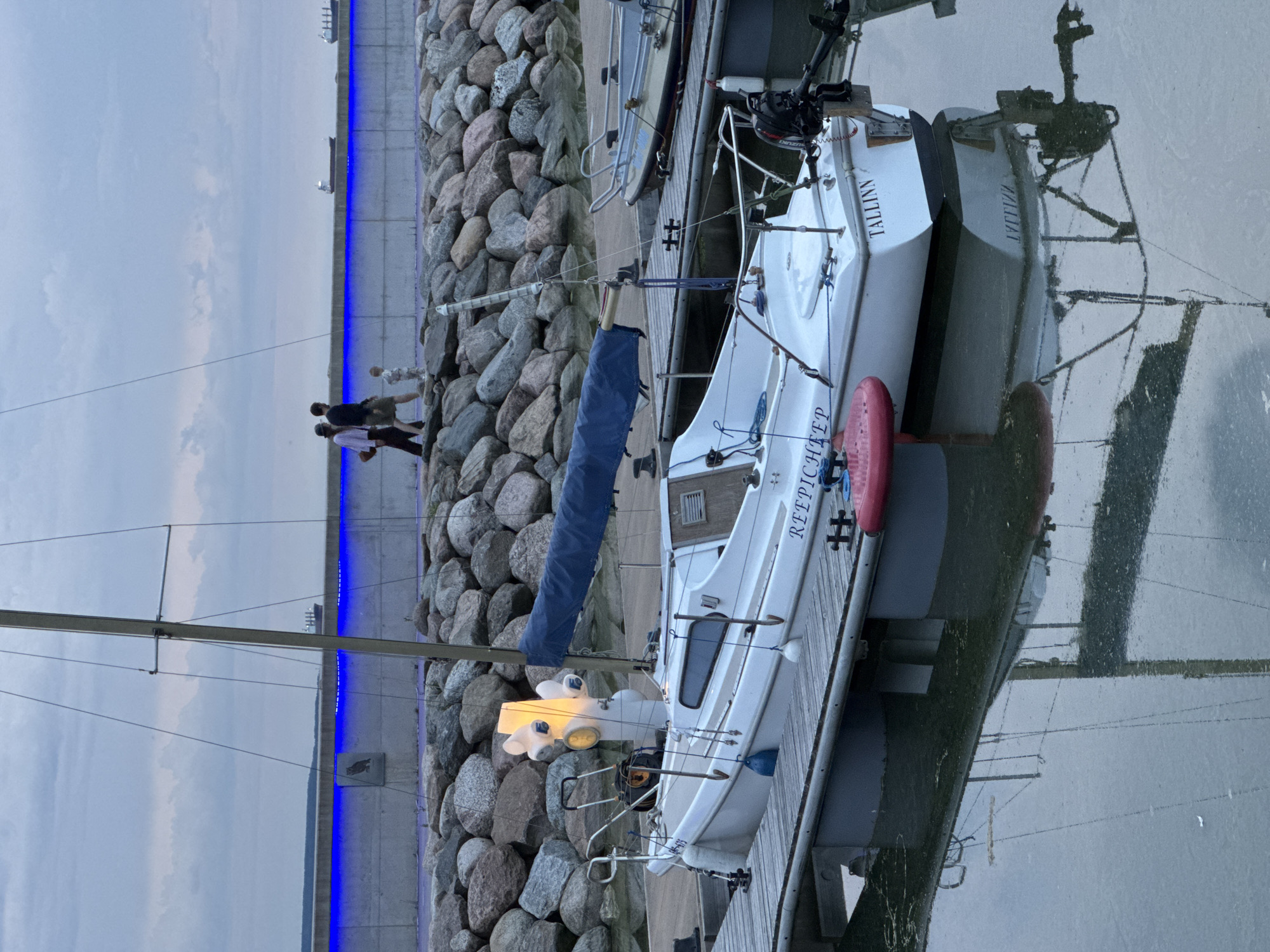 A sailboat named "TALLINN" is docked at a pier in Tallinn, Estonia. The sailboat is white with a blue sail cover. It has a small outboard motor on the back. There is a red buoy attached to the side of the boat. In the background, a breakwater is illuminated with blue lights. People are walking along the top of the breakwater, and ships are visible in the distance. The water is calm and reflects the sailboat and the sky.