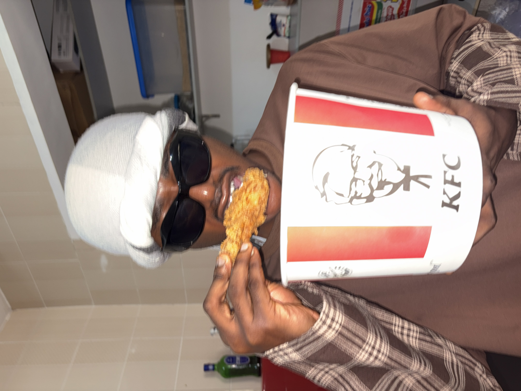 The image features a person holding a bucket of KFC fried chicken and a piece of chicken. The person is wearing a white beanie, sunglasses, and a brown plaid shirt over a brown long-sleeved shirt. They are in the process of eating a piece of fried chicken. The background appears to be an indoor setting, possibly a kitchen or a storage area, with shelves and some containers visible. A green bottle is also visible on a surface in the lower part of the image. The lighting suggests it is daytime or a well-lit indoor environment. The person's expression is somewhat obscured by the sunglasses, but the overall impression is casual and relaxed, enjoying a meal. The KFC bucket has the recognizable logo and branding. There is no indication of the time of day or weather conditions. The location context is Igboefon, Nigeria.