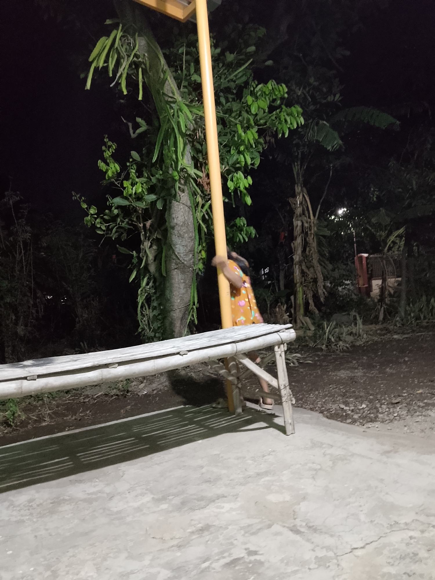 A young person wearing an orange and yellow patterned dress is leaning against a bright yellow pole in Kelurahan, Indonesia at night. The pole is located near a bamboo bench with a weathered gray top and white legs, which casts a striped shadow on the concrete ground. The background is dark with lush vegetation, including a tree with hanging epiphytic plants, and appears to be a rural or semi-rural outdoor setting. There is a hint of a building with red accents in the distance. The lighting suggests artificial illumination from the yellow pole itself or a nearby light source.