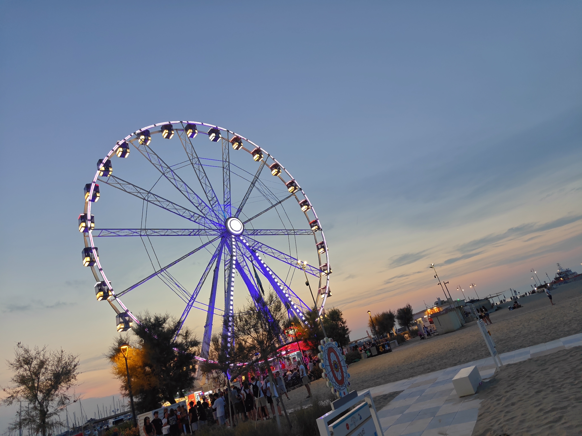 The image depicts a Ferris wheel illuminated with purple and white lights at dusk on a beach in Rimini, Italy. The Ferris wheel is the central subject, with its cabins visible as squares with yellow lights. The sky is a gradient of blue and orange, indicating the sunset.
In the foreground, there is a sandy beach with a checkered tiled pathway. Several people are gathered around the base of the Ferris wheel and near various kiosks and stalls. These structures appear to be part of a fairground or amusement area set up on the beach. Some trees are also visible, casting shadows on the sand.
In the background, the sea is visible with a few boats docked. The overall atmosphere suggests a lively evening on the Rimini coast. The image is taken from a slightly elevated angle, looking towards the Ferris wheel and the beach.