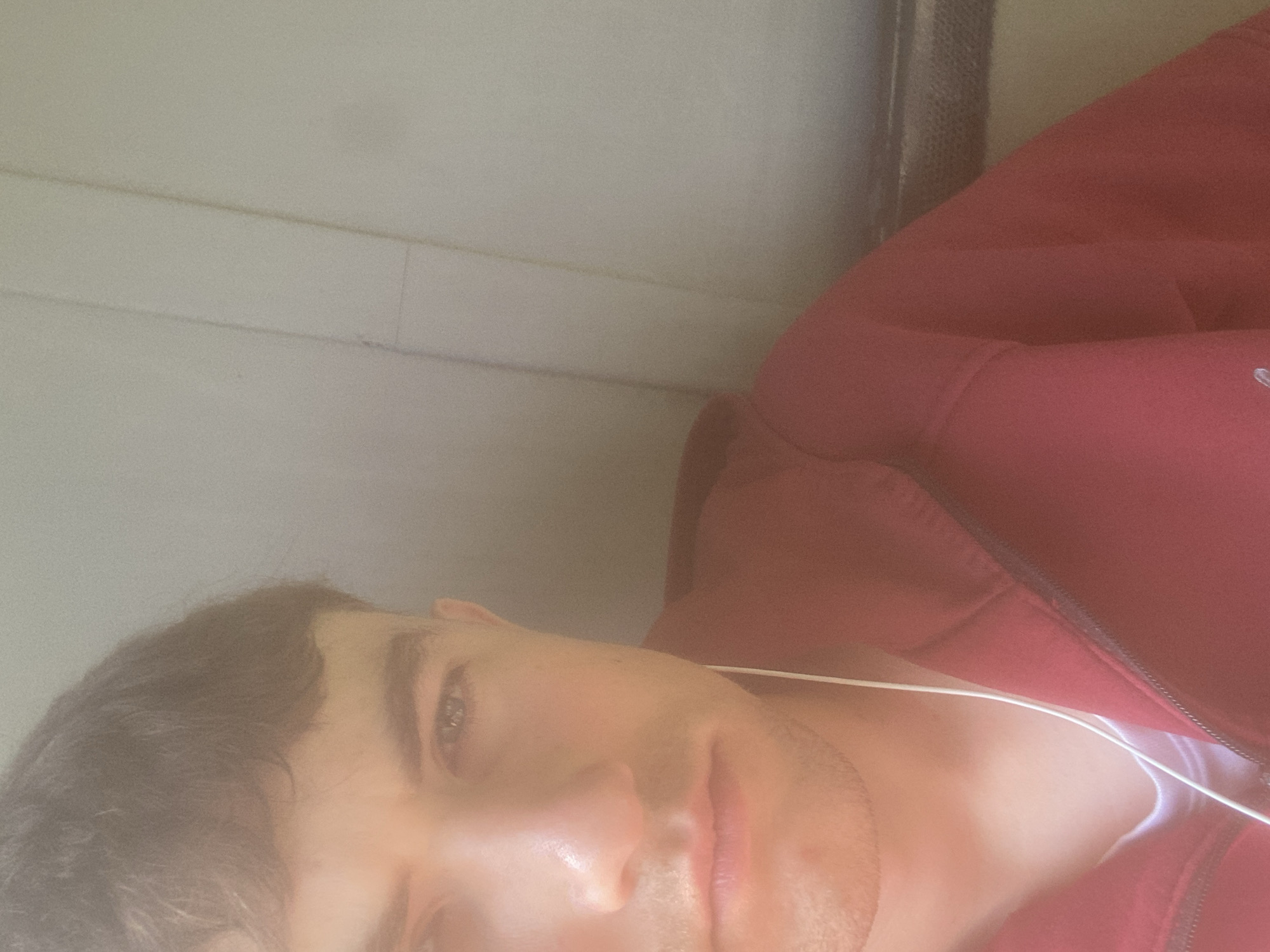 A close-up, rotated view shows a person's face partially in frame. They have short dark hair, light skin, and are wearing headphones. They are wearing a red zippered jacket. The background consists of a light-colored surface with horizontal lines, possibly a wall. The photo's origin in Matyashivka, Ukraine is not determinable from the image content.