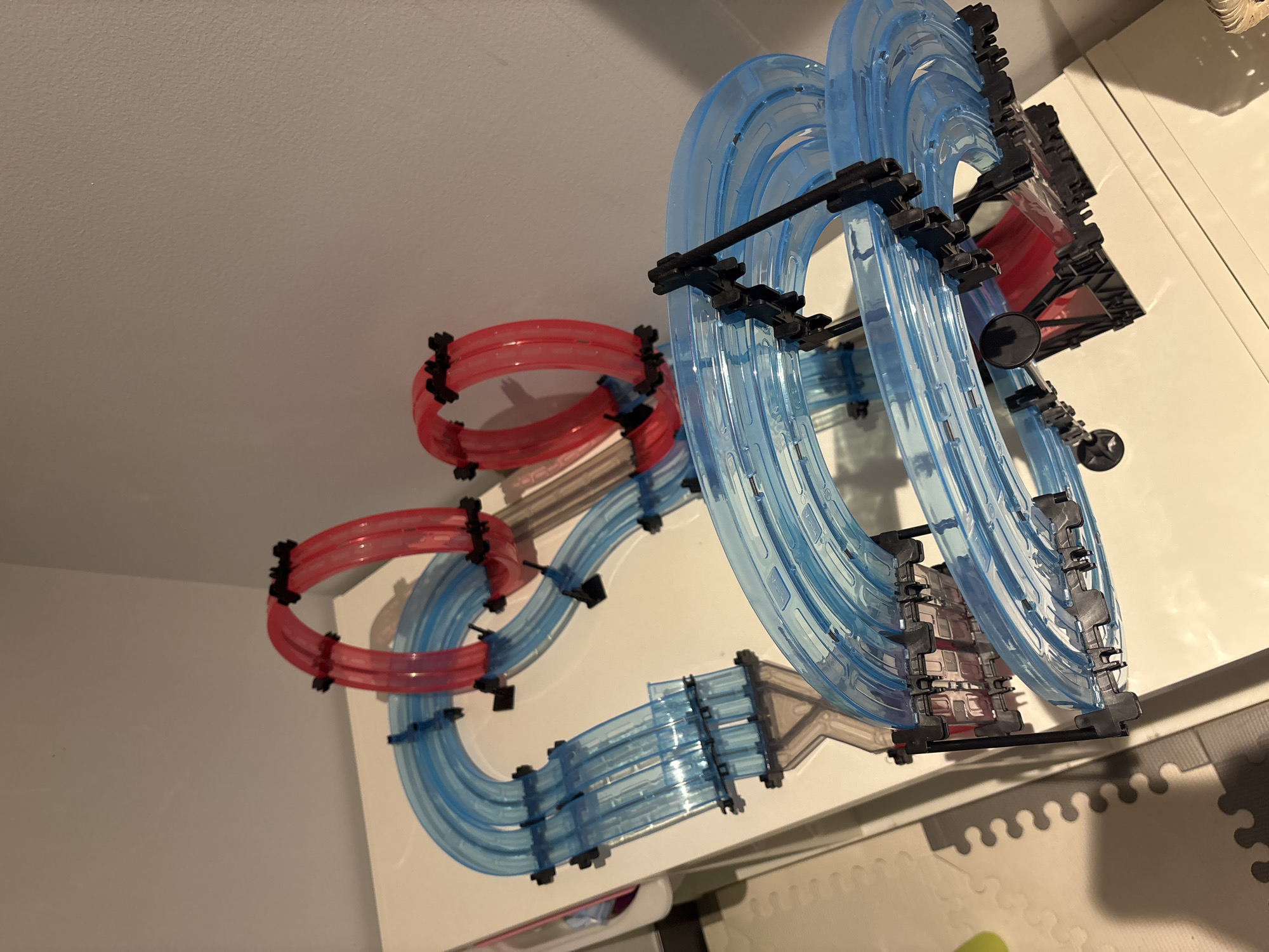 An overhead, slightly angled, medium shot shows a toy race track on a white table against a light gray wall. The race track is made of clear blue plastic tubes with black connectors and red plastic tube loops. The blue tubes are arranged in curves and straight sections. The red loops are vertical and are spaced apart. A portion of a patterned floor mat is visible in the lower left corner. There are no people present. The scene is indoors.