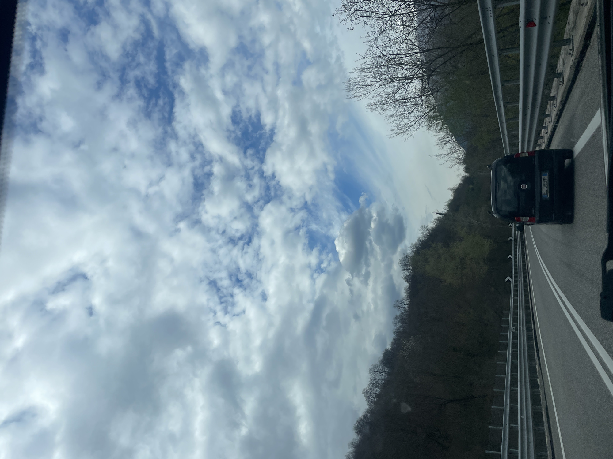 A photograph taken from within a moving vehicle shows an upward view of a partly cloudy sky. The clouds are mostly white and gray, with patches of blue sky visible. A dark-colored Fiat Qubo vehicle is driving on a divided highway. A metal guardrail runs parallel to the road, and a wooded area lines the edge of the highway. The vehicle has a license plate with the prefix EL, and the scene appears to be taken in Collepiano, Italy, based on the visible license plate and likely surroundings.