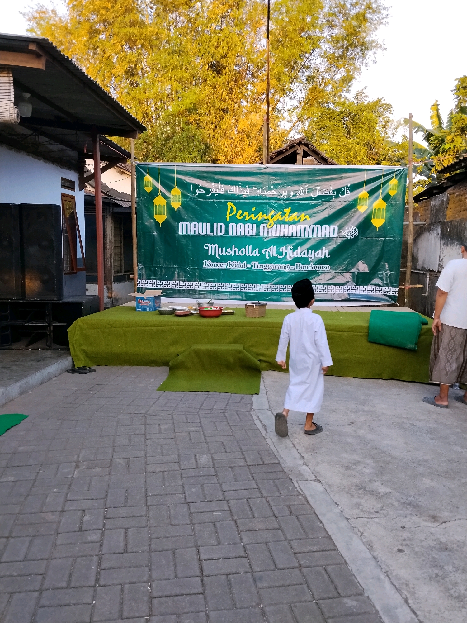 An outdoor daytime scene in the city of Kejawan, Indonesia, captures a public event. A large green banner with yellow lanterns and Arabic calligraphy hangs prominently, indicating a celebration for "Maulid Nabi Muhammad" at "Musholla Al-Hidayah," located in Kidul-Tenggarang-Bondowoso. The banner is displayed on a wooden frame. In front of the banner, a green, carpeted platform is set up, adorned with several bowls, plates, and a red bowl, suggesting a meal service or offerings. To the left of the platform, two large black speakers are positioned. The ground is paved with interlocking grey bricks. A young boy, dressed in a white robe and a black cap, walks away from the camera across the paved area. To the right, an adult male, wearing a white shirt and a patterned sarong, stands facing away from the camera. The background features bamboo trees with yellowing leaves and parts of buildings with corrugated metal roofs. The overall atmosphere appears to be festive and communal.