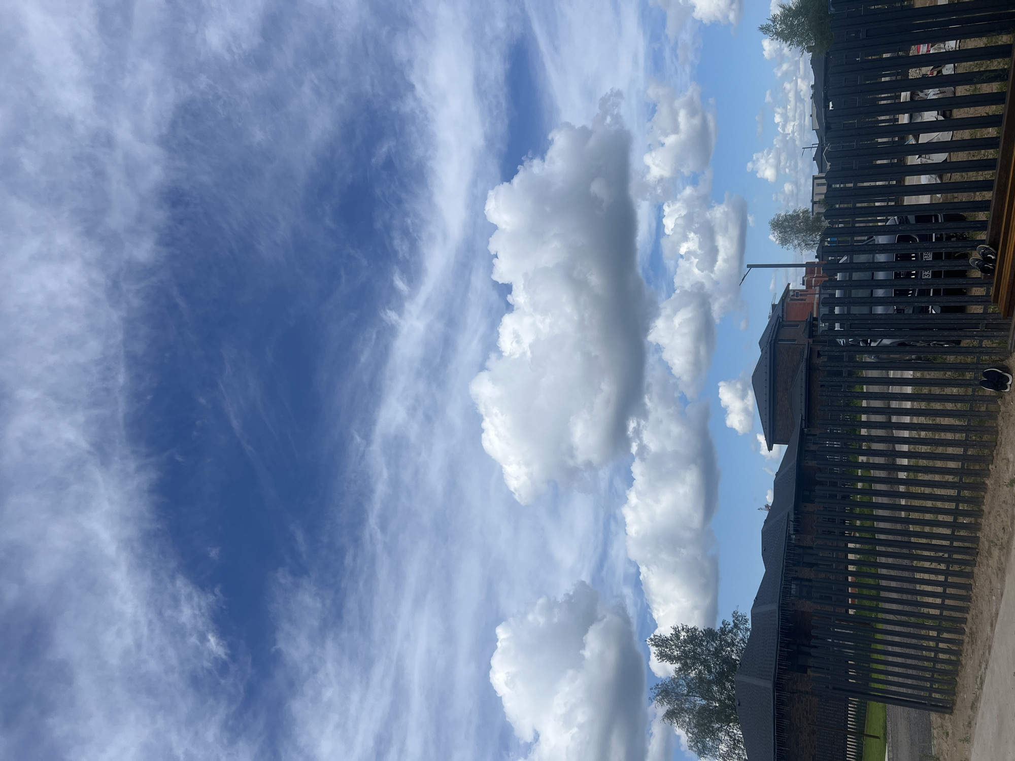 In Yurlovo, Russia, a dark metal fence runs horizontally across the foreground, obscuring a view of several modern brick houses with dark roofs. Behind the fence, a black SUV is parked. The sky above is a bright blue, filled with a mix of wispy cirrus clouds and large, puffy cumulus clouds. A few trees are visible, some with sparse foliage, suggesting it is either early spring or late autumn. The ground in front of the fence is sandy, with some construction materials, including metal beams, visible on the right side. A pair of black and white sneakers and a pair of black flip-flops are placed near the bottom right corner of the image.