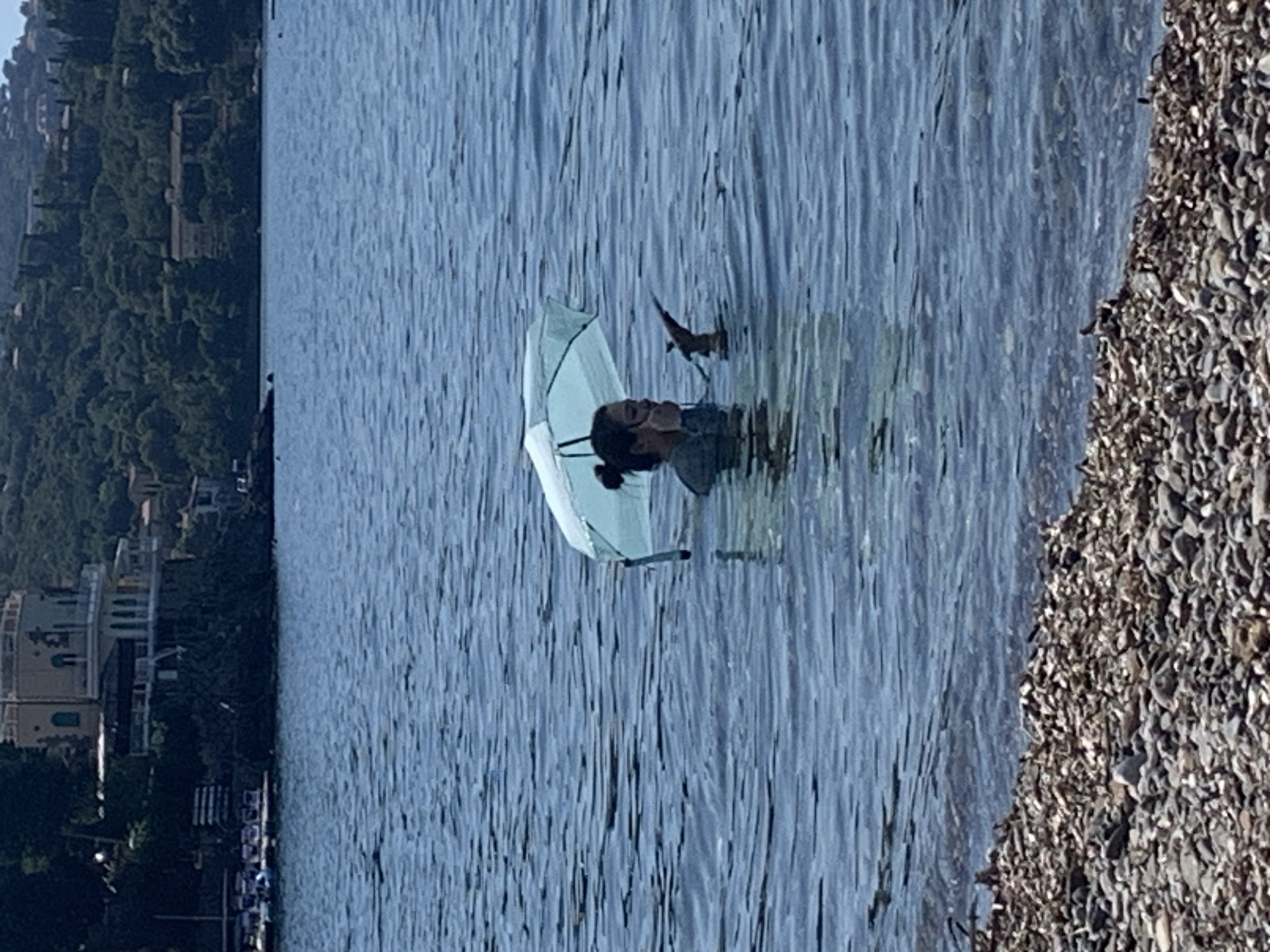 In the city of Pozzarello, Italy, a person stands in the water holding a light blue umbrella. The person is submerged up to their chest and appears to be holding a phone or similar device in their right hand. The background features a shoreline composed of pebbles, with a body of water extending towards a distant coastline characterized by dense green foliage and buildings. The sky is clear and blue, suggesting a sunny day.