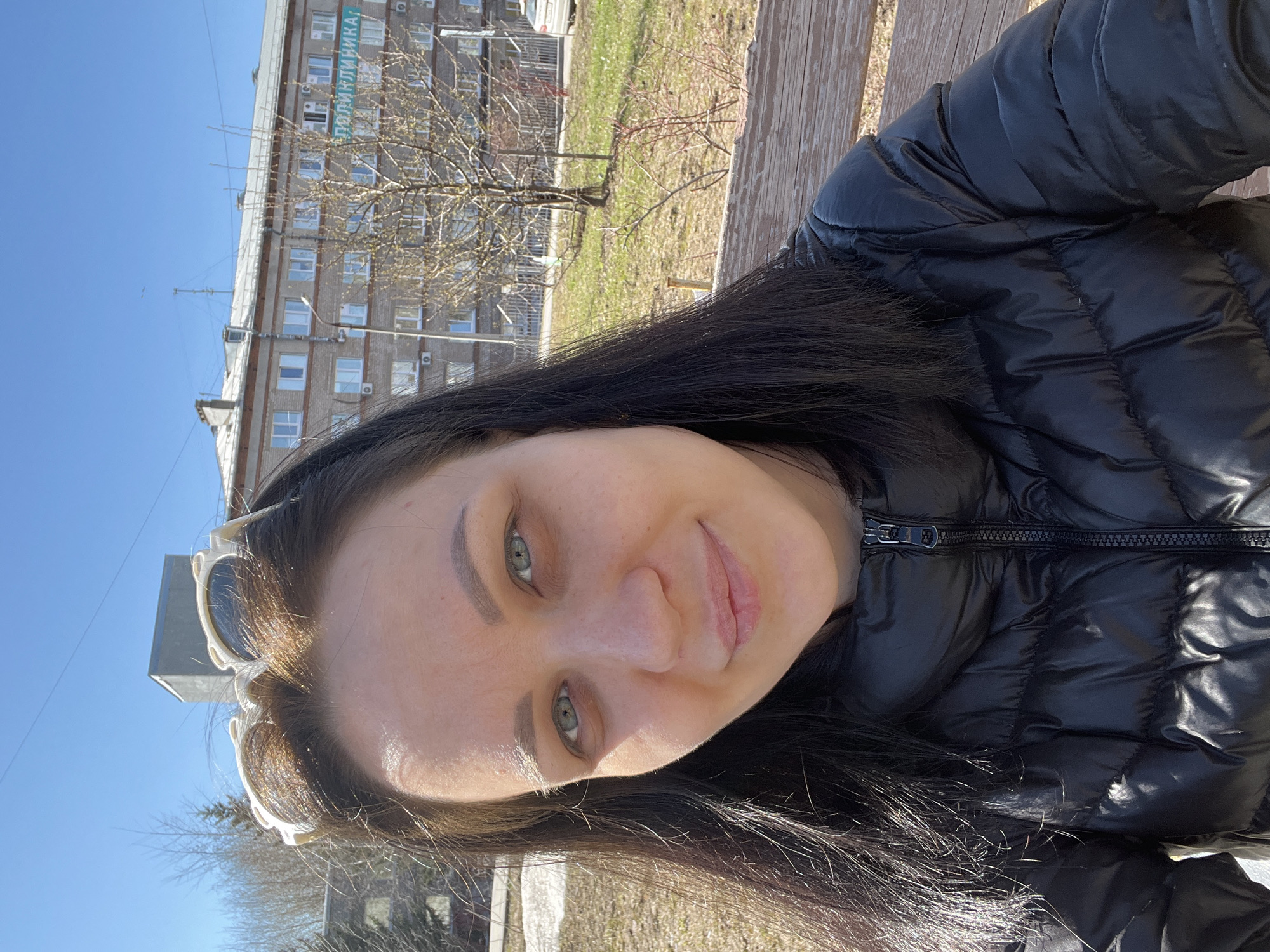 A woman with long dark hair and light eyes, wearing a black puffy jacket, is photographed outdoors. The location appears to be a city park in Perm, Russia, given the presence of a building with a sign that includes the Cyrillic letters "ПОЛИКЛИНИКА," which translates to "Polyclinic". The woman is likely seated on a wooden bench. She is looking directly at the camera with a slight smile. Sunglasses are positioned on top of her head. The background shows a mix of green grass, bare tree branches, and a clear blue sky.