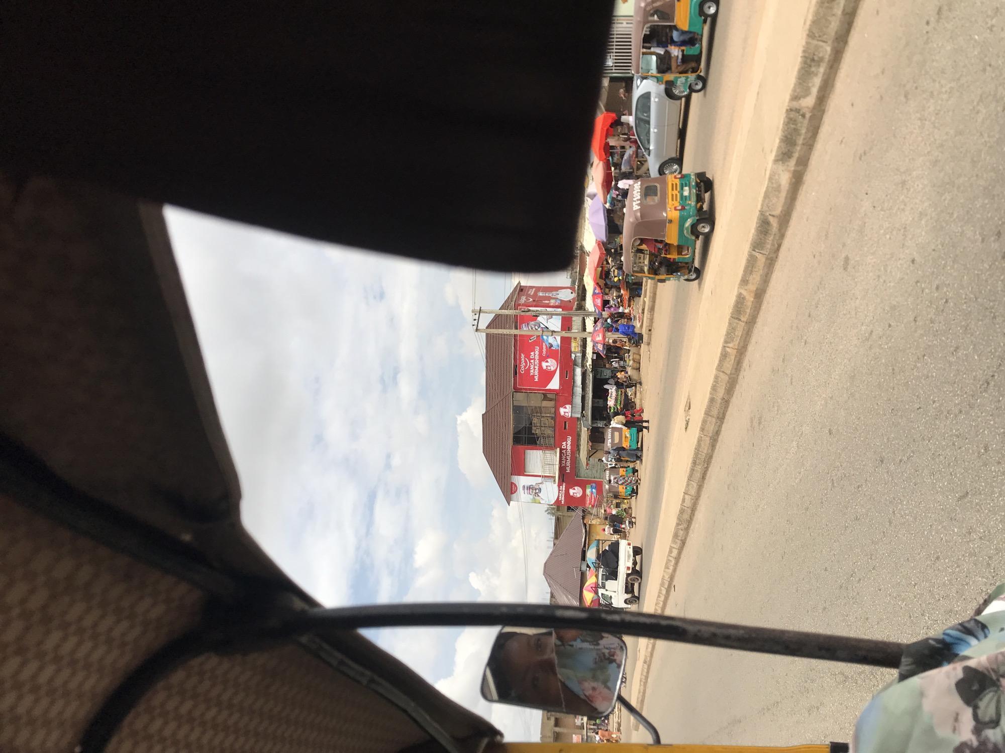 The image captures a bustling street scene in Kabong, Nigeria, observed from inside a vehicle, likely a tricycle, with its roof and internal frame partially visible. The immediate foreground shows the vehicle's frame and a side mirror reflecting the face of an individual wearing a patterned face mask.
The asphalt road, lined with a concrete curb, extends from the foreground into the mid-ground, where various vehicles and pedestrians are present. On the left side of the road, a white pick-up truck is in motion, followed by several people. A light green, yellow, and brown tricycle (keke napep) with license plate "PT 183U2" is on the road, alongside a silver sedan, and another similar tricycle further right.
Beyond the road, a vibrant commercial area unfolds. A prominent two-story building with a brown roof dominates the center-right, its lower facade painted red and adorned with large "Colgate" advertisements featuring a smiling man and the text "YANGA DA MURMUSHINKU". In front of this building and extending to the left, numerous market stalls are set up, shaded by colorful umbrellas in red, purple, and other hues. Many individuals are visible around these stalls, engaging in market activities, walking, or standing.
The sky is overcast with a mix of white and gray clouds. Utility poles and overhead wires are visible against the sky to the left of the large red building. The scene depicts a typical active market environment in a Nigerian urban setting.
