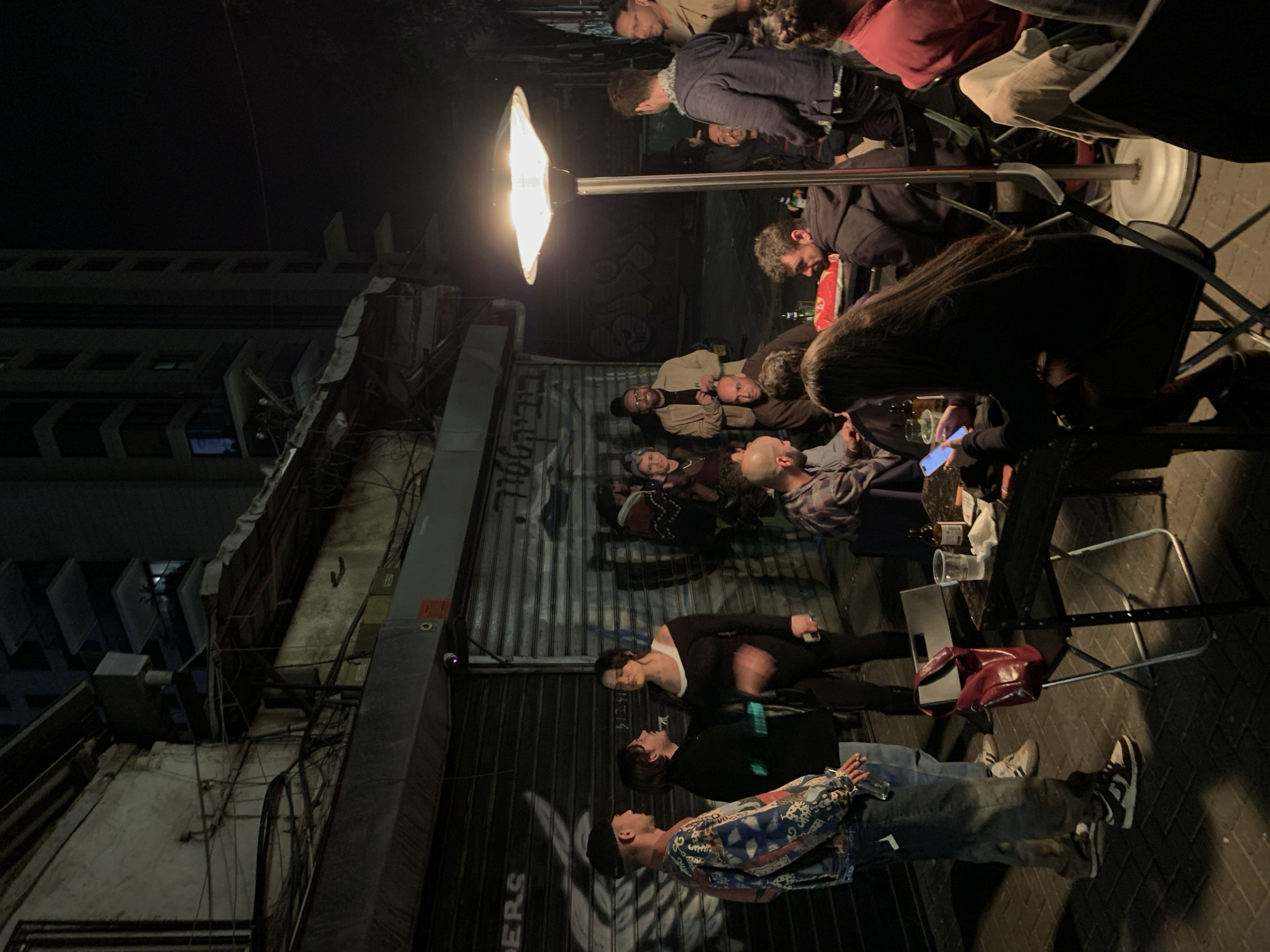 People are gathered at an outdoor bar in Tel Aviv, Israel. They are sitting at tables and talking. A bright light illuminates the scene. The building behind the bar is old and dilapidated. The bar is on a narrow street in a busy area of the city. The image shows various individuals in casual clothing, some seated at tables with drinks, others standing and interacting. There are several chairs and tables scattered around the scene, indicating the bar's outdoor seating area. The lighting is dominated by a single bright light source. The backdrop includes a corrugated metal door with graffiti. The setting appears to be an urban environment with older buildings.