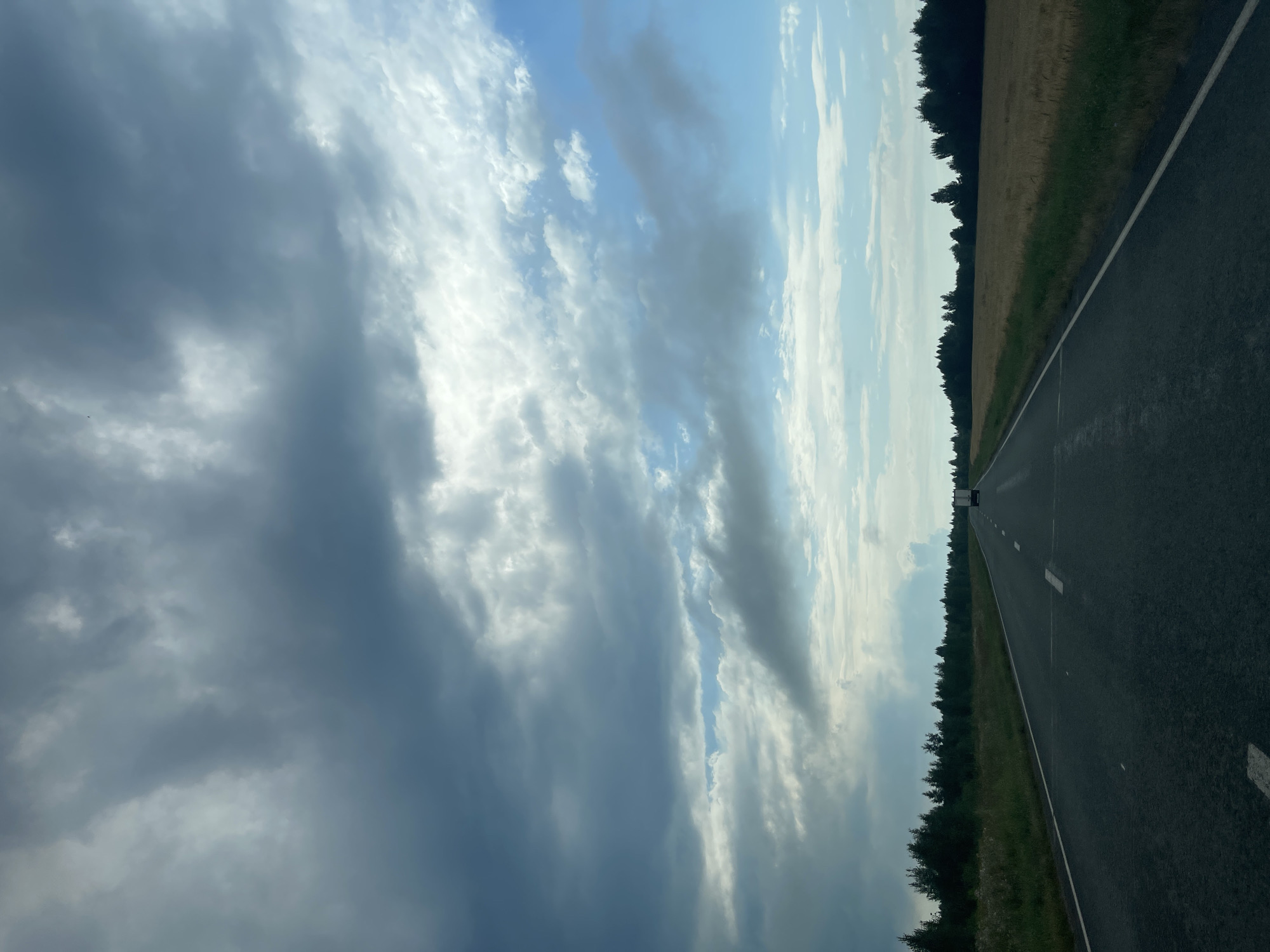 The image depicts a road stretching into the distance, flanked by fields and trees. Dark, heavy clouds dominate the sky, with patches of blue sky visible. A truck is visible in the distance on the road. The location is identified as Sidorovy Gory, Russia. The scene is a view from a vehicle traveling on a paved road through a rural landscape under a cloudy sky.