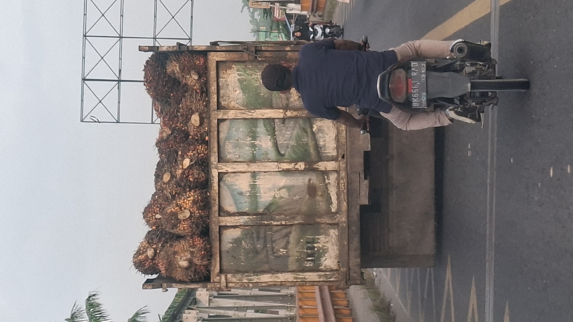 In Tanjung Pura, Indonesia, a motorcyclist wearing a dark blue shirt and tan pants rides behind a large truck filled with harvested palm oil bunches. The truck's side panels are painted with faded murals depicting mountainous landscapes. The sky is overcast, and the road appears to be asphalt with yellow lane markings. In the distance, another motorcyclist with a passenger is visible on the same road. The scene is captured from a slightly elevated rear perspective of the leading motorcycle.